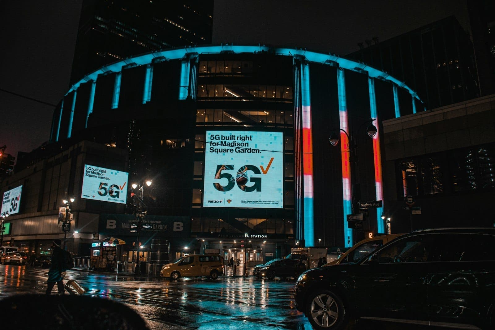 ด้านนอกของ Madison Square Garden ในยามค่ำคืน พร้อมไฟนีออนหลากสี ป้ายดิจิทัล ถนนเปียก และรถยนต์ใน Midtown Manhattan