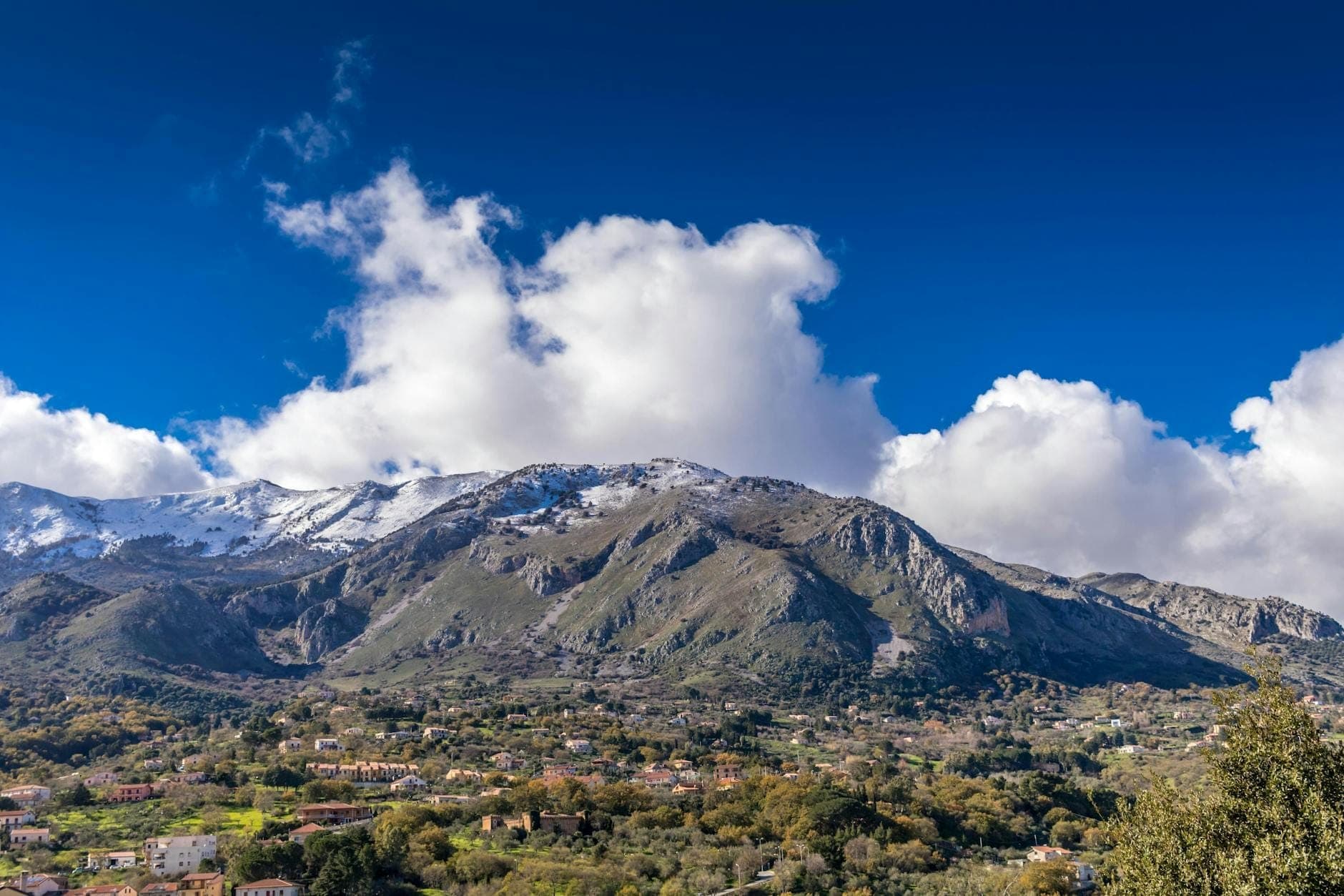 Pemandangan panorama pegunungan terjal dengan puncak bersalju, lembah hijau, kota-kota abad pertengahan yang tersebar, dan awan dramatis di bawah langit biru cerah di pedalaman Sisilia.