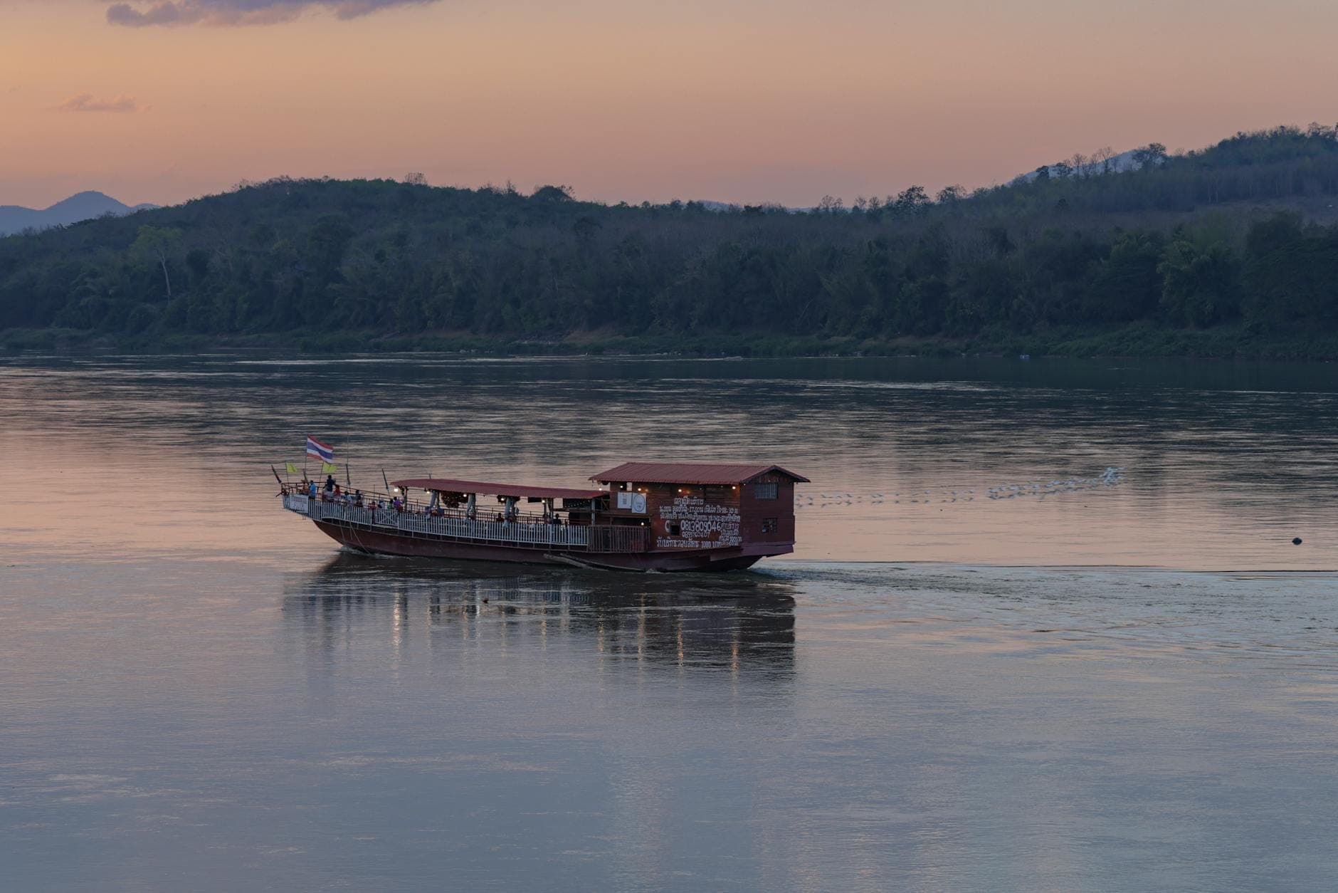 Ein traditionelles Longtail-Boot mit überdachtem Deck gleitet bei Sonnenuntergang einen ruhigen Fluss entlang, im Hintergrund bewaldete Hügel.