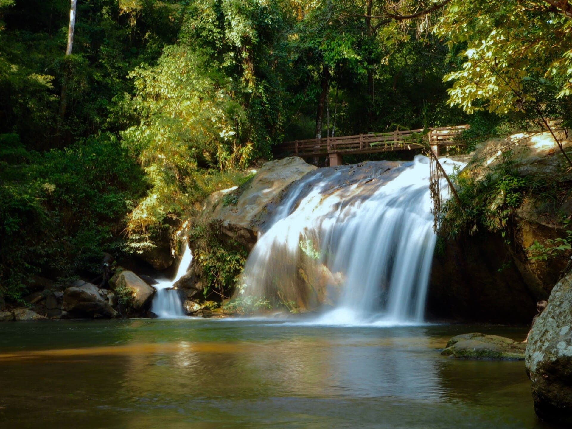 La cascada Mae Sa cae sobre grandes rocas bajo un puente de madera, rodeada de un frondoso bosque verde en el valle de Mae Rim.
