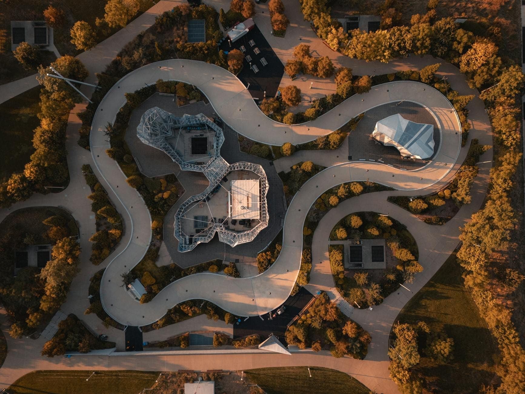 Aerial view of Maggie Daley Park showing winding paved paths, geometric playground structures, lush green trees, and golden sunlight over landscaped grounds.