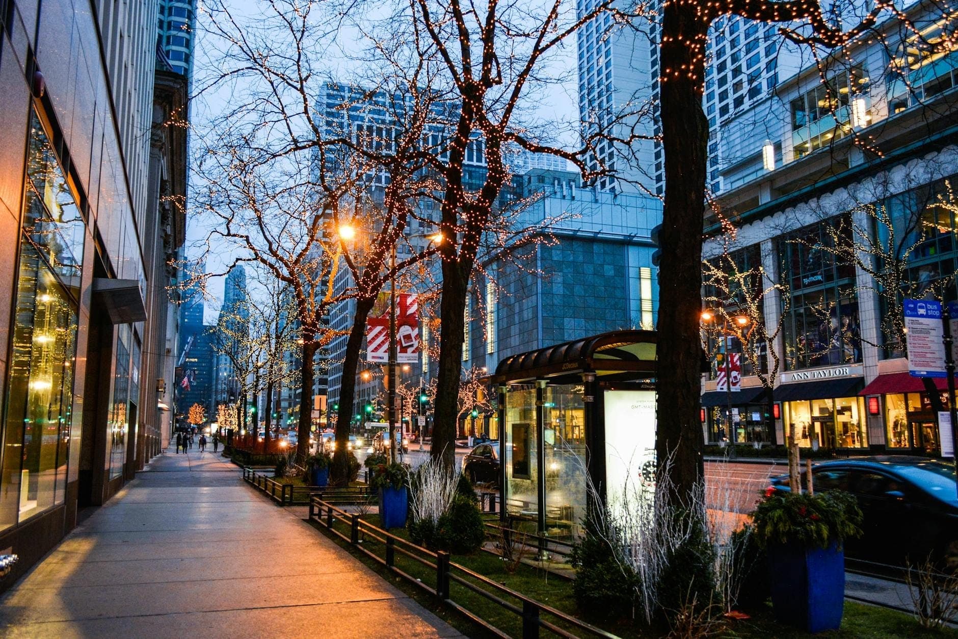 Evening street view of Chicago's Magnificent Mile with twinkling lights in trees, luxury storefronts, and modern skyscrapers lining the bustling sidewalk.