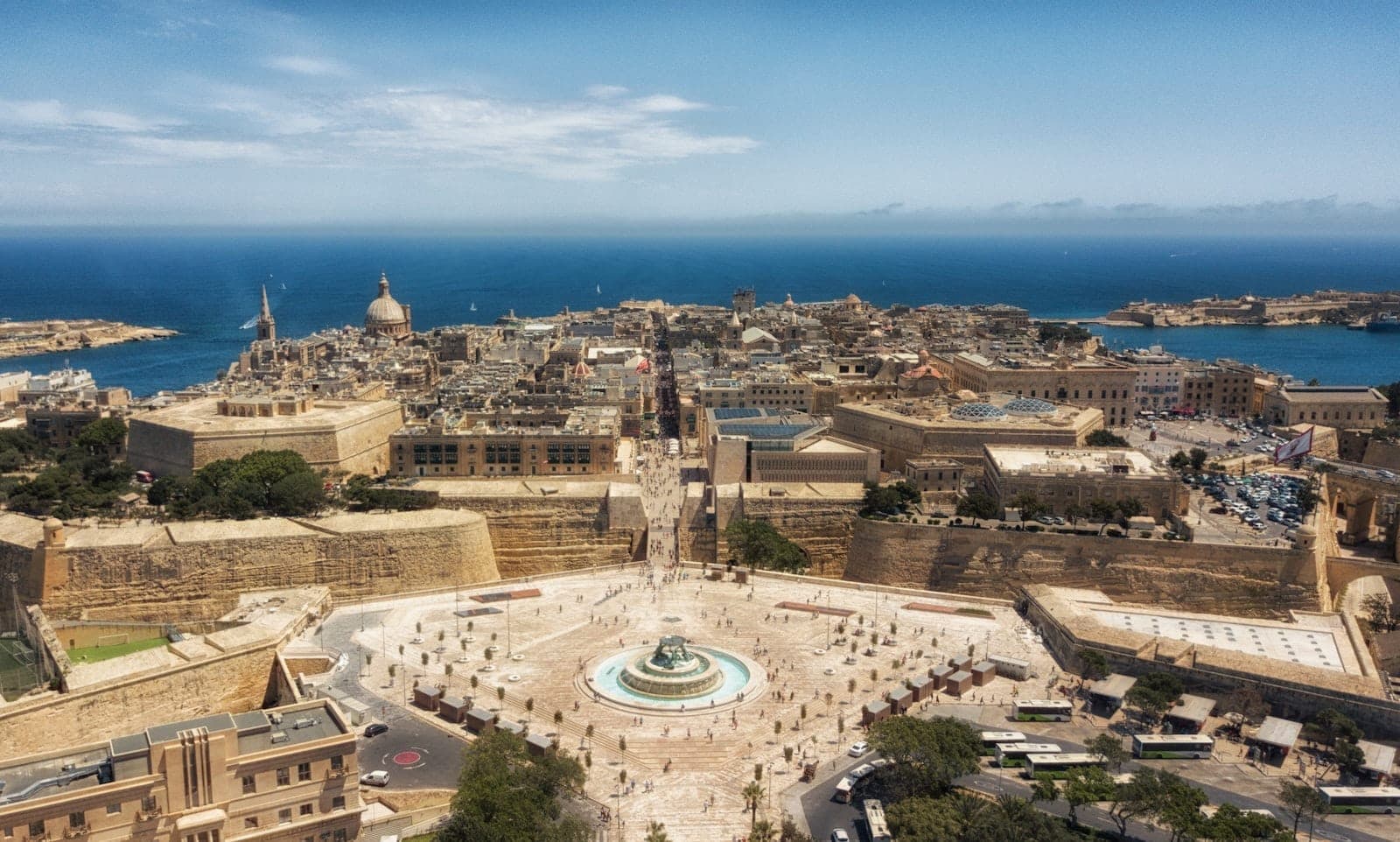 Panoramischer Luftblick über Valletta, Malta, mit historischen Kalksteingebäuden, einem großen Kreisverkehr mit Brunnen und dem Mittelmeer im Hintergrund unter blauem Himmel.