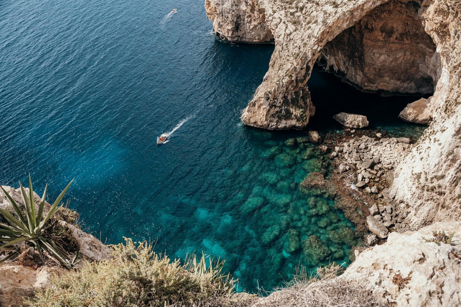 Vue aérienne du littoral escarpé de Malte avec un imposant arc de grotte marine naturelle, une eau turquoise cristalline et un petit bateau à proximité.