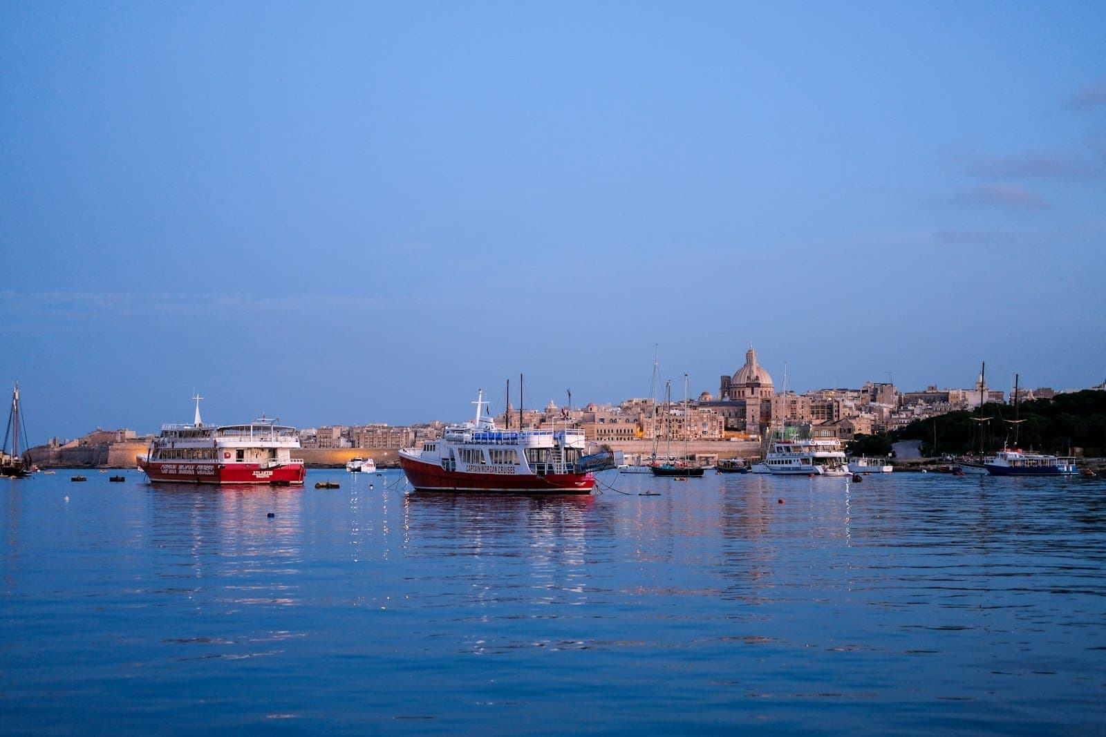 Bateaux flottant sur des eaux calmes près d'un front de mer maltais historique au crépuscule, avec des bâtiments de la ville en arrière-plan.