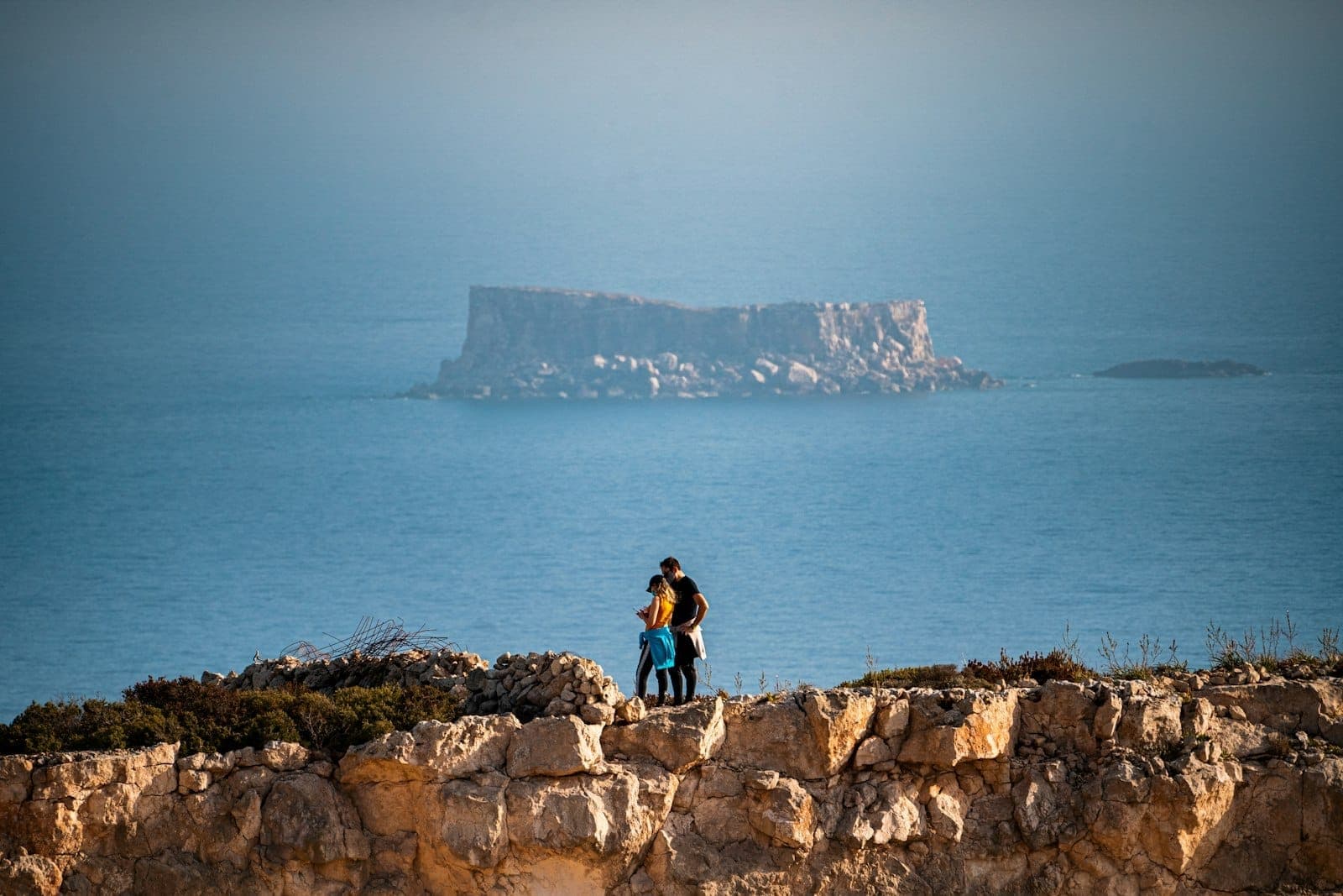 Ein Paar steht eng beieinander auf einer Klippe und blickt aufs Meer, im Hintergrund eine felsige Insel unter klarem Himmel in Malta.
