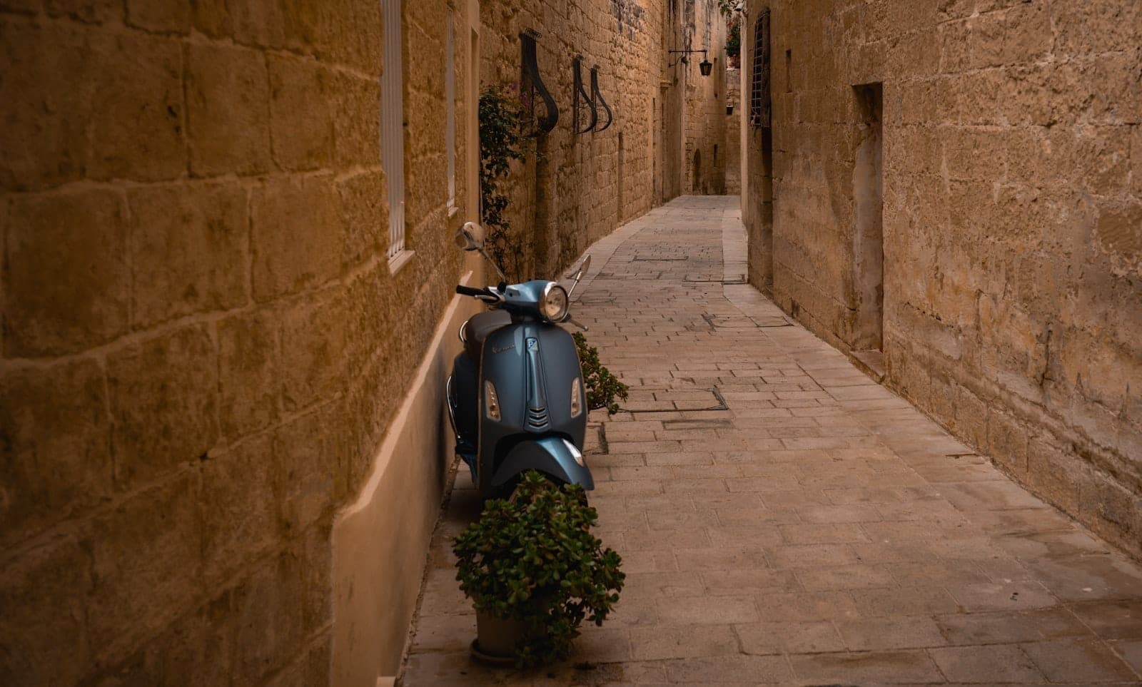 Eine schmale Steingasse in Mdina, Malta, mit einem blauen Roller, der neben alten Kalksteinmauern und Topfpflanzen geparkt ist.