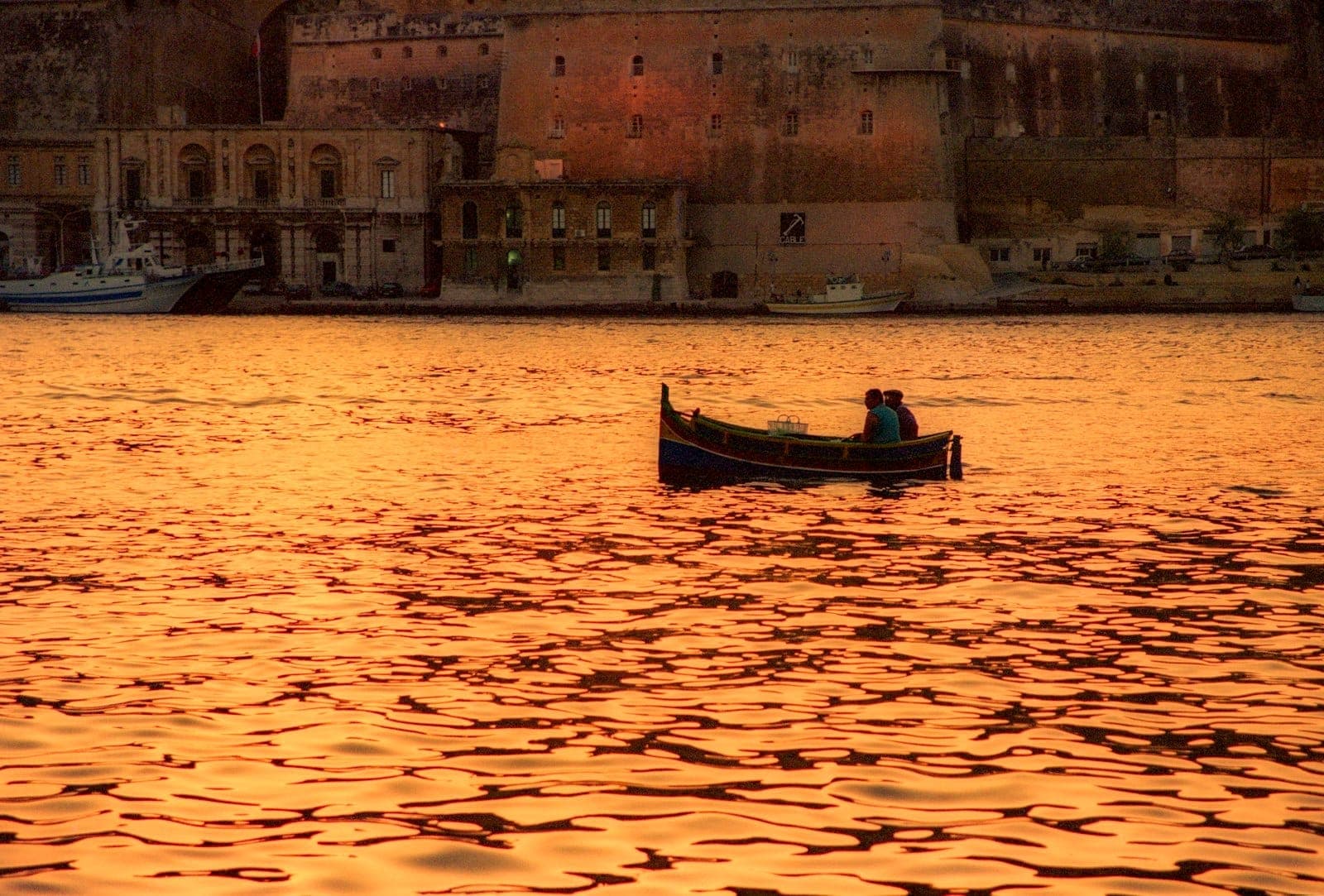 Kleines Boot mit einem Pärchen an Bord bei Sonnenuntergang, mit goldenem Wasser und historischen Steingebäuden von Malta im Hintergrund.