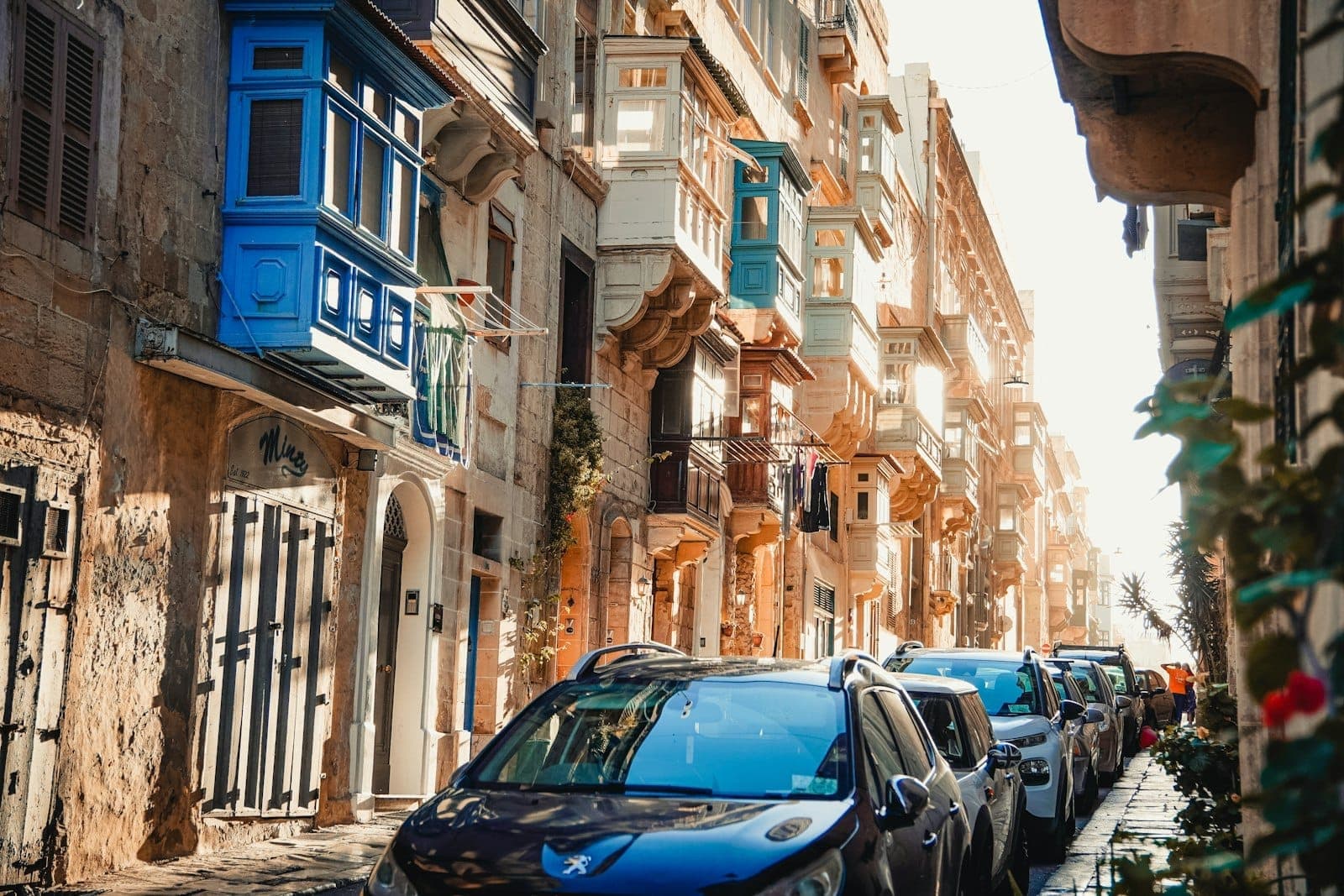 Sonnenbeschienene Straße in Valletta, gesäumt von honigfarbenen Barockgebäuden und traditionellen maltesischen Balkonen, Autos entlang der schmalen Straße geparkt.