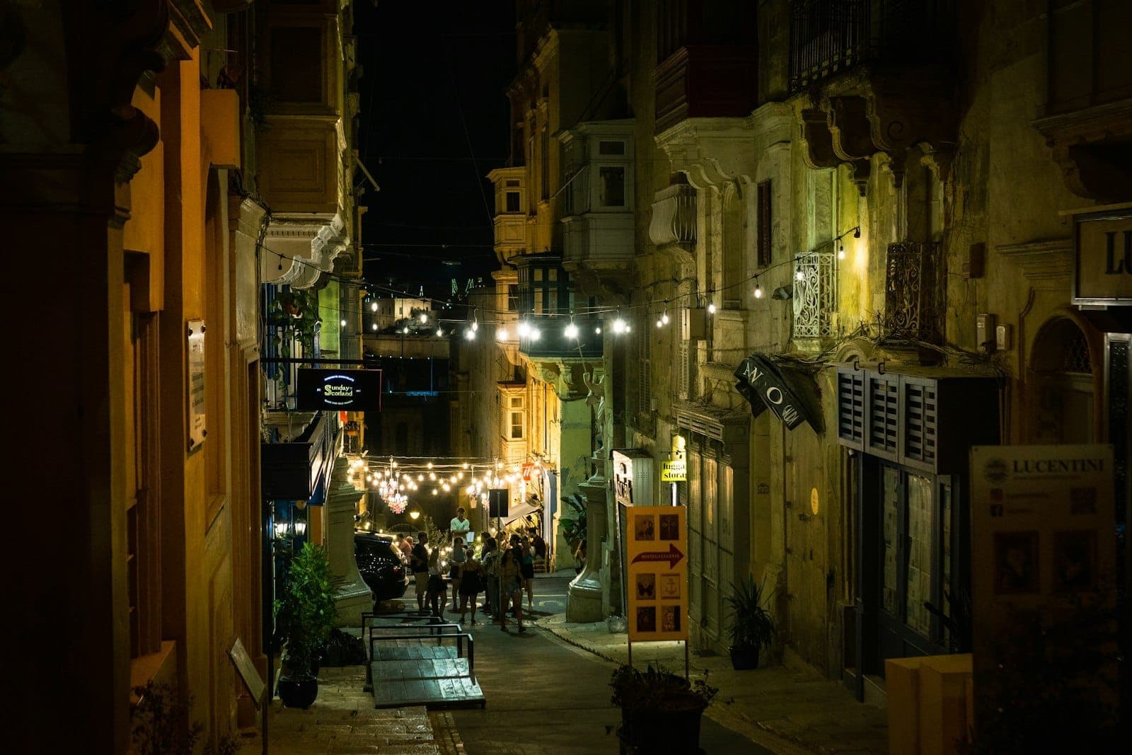 Enge Gasse in Valletta bei Nacht mit leuchtenden Lichterketten, Menschengruppen vor Bars und historischen Gebäuden auf beiden Seiten.