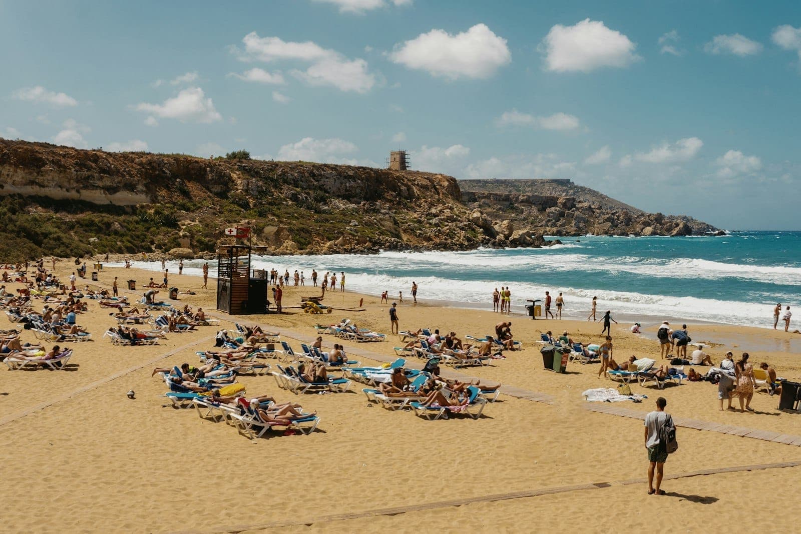 Beliebter Sandstrand auf Malta an einem sonnigen Tag, mit vielen Menschen beim Sonnenbaden und Schwimmen und einem felsigen Vorgebirge im Hintergrund.