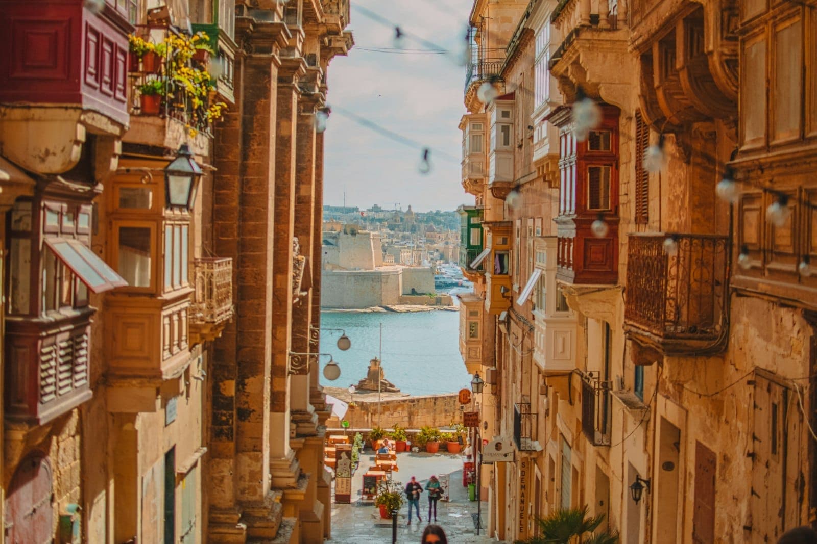 Sonnige maltesische Straße, die zum Wasser hinunterführt, gesäumt von historischen Steingebäuden mit Balkonen – eine einladende und sichere Reiseatmosphäre.