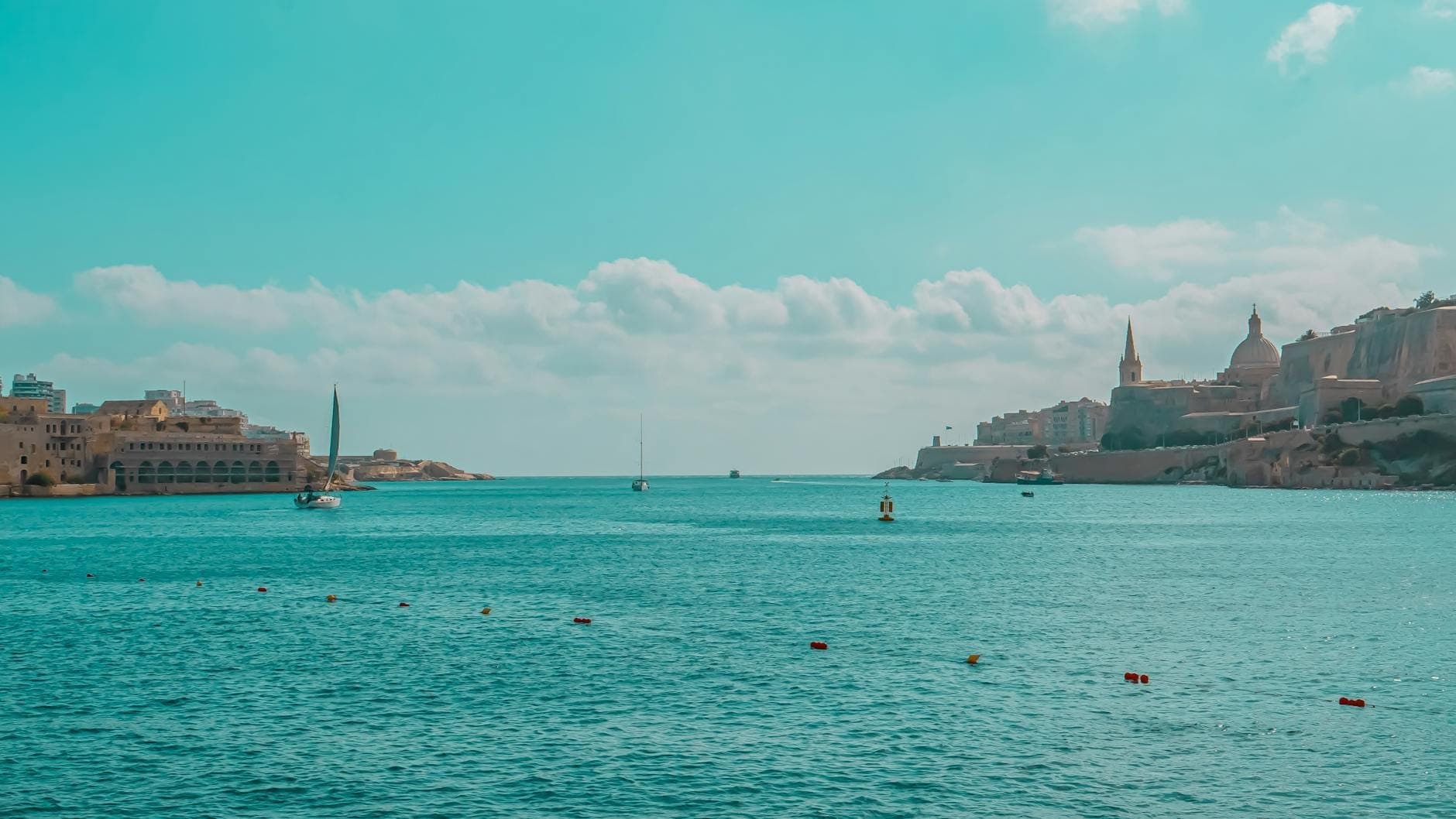 Ruhiges türkisfarbenes Meer mit Segelbooten und der historischen Skyline Vallettas unter blauem Himmel – der malerische Abfahrtspunkt der Fähre nach Sizilien.