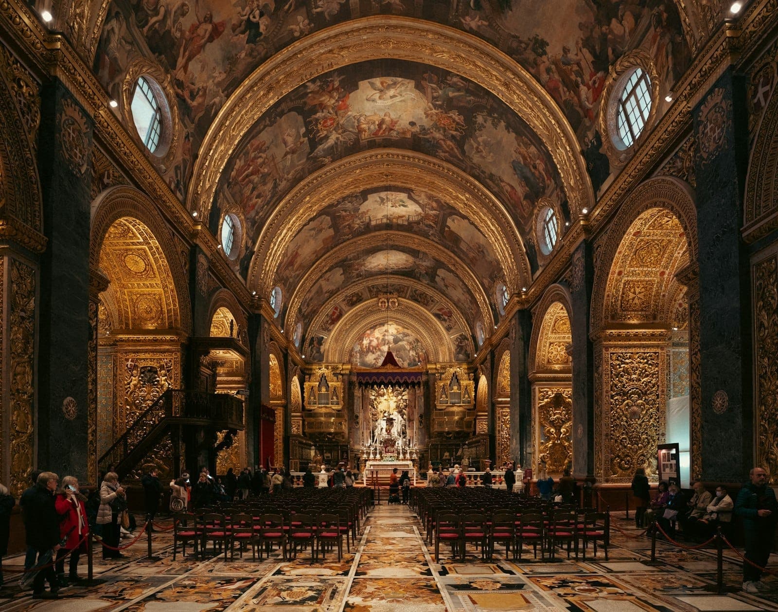 Interno ornato di una chiesa maltese a Valletta, con archi dorati, affreschi dettagliati e visitatori seduti e in piedi all'interno.