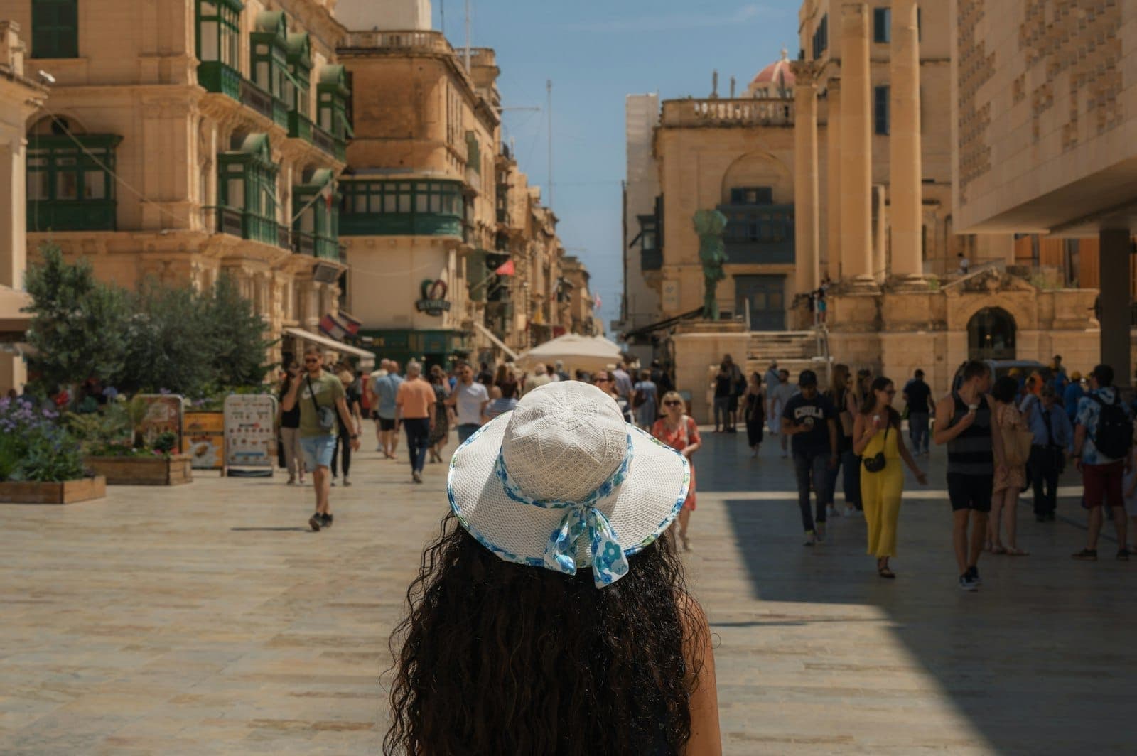 Vista di una soleggiata strada maltese fiancheggiata da edifici storici, animata da pedoni e una persona con cappello da sole in primo piano.