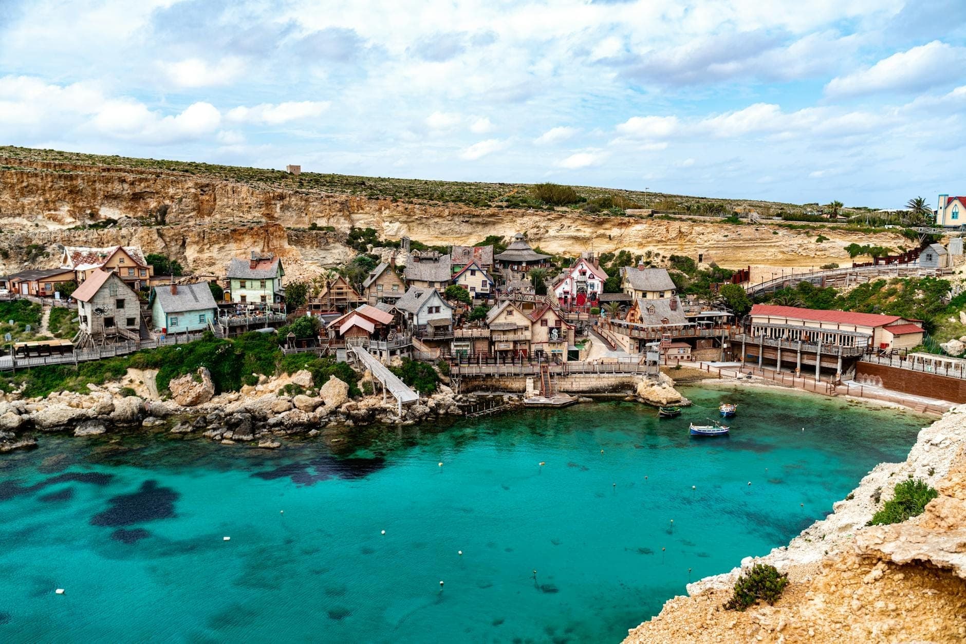 Vista ampia di Popeye Village con colorati edifici in legno sulla turchese Anchor Bay a Malta sotto un cielo azzurro.