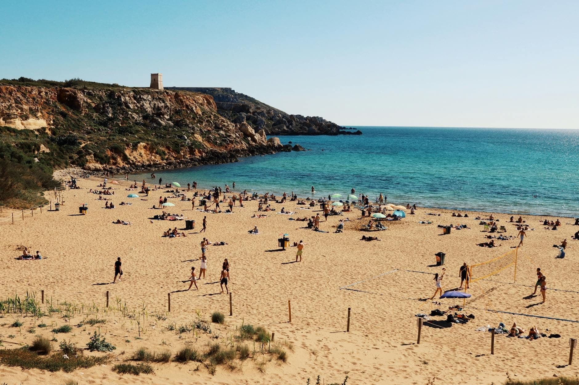 Un'ampia spiaggia di sabbia a Malta con famiglie e bambini che giocano, bagnanti che prendono il sole e acque blu poco profonde. Sullo sfondo sono visibili scogliere rocciose e una torre di avvistamento.