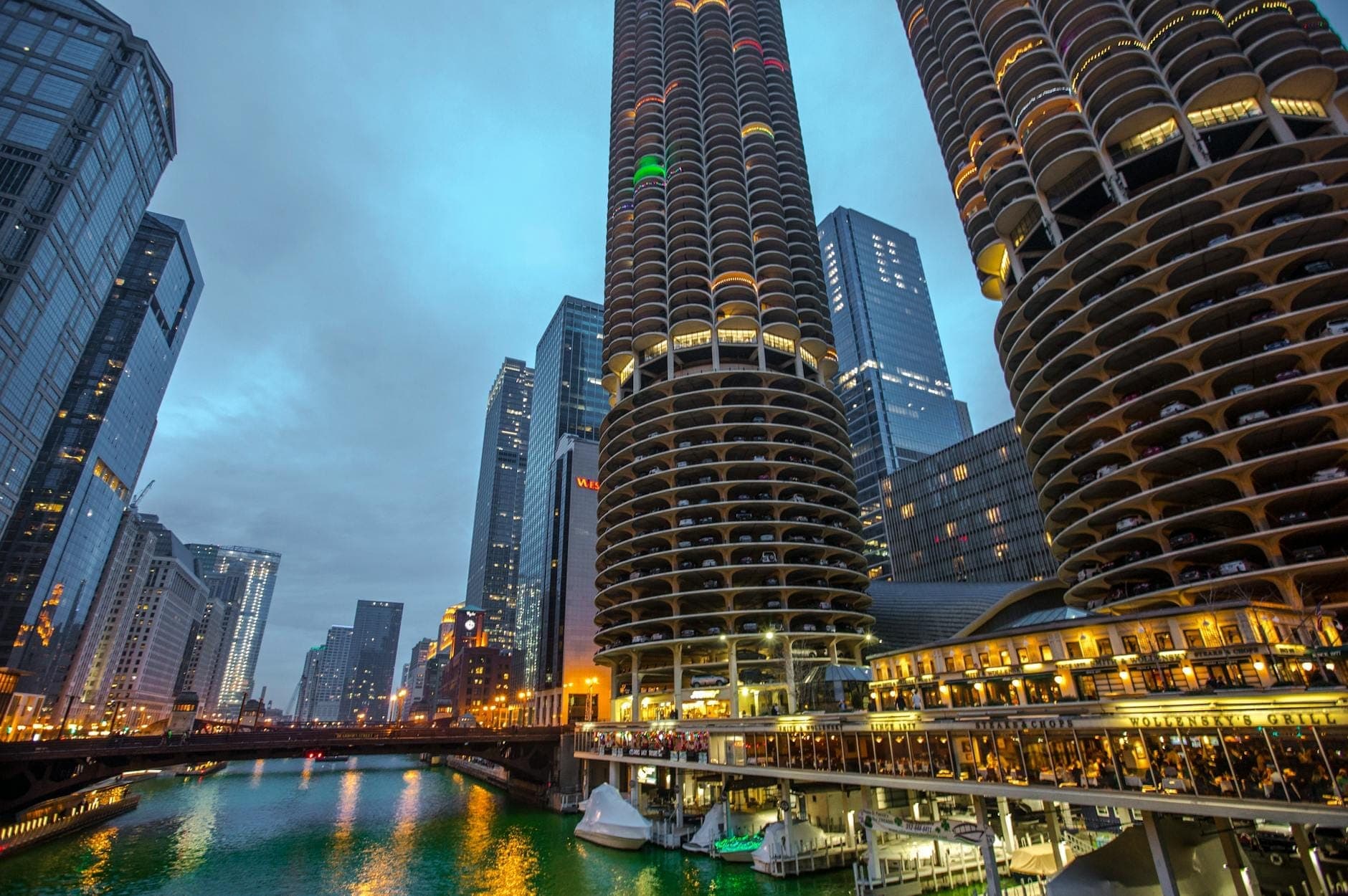 Evening view of Marina City’s two cylindrical towers on the Chicago River, surrounded by skyscrapers, city lights reflected in the water, and riverwalk activity.