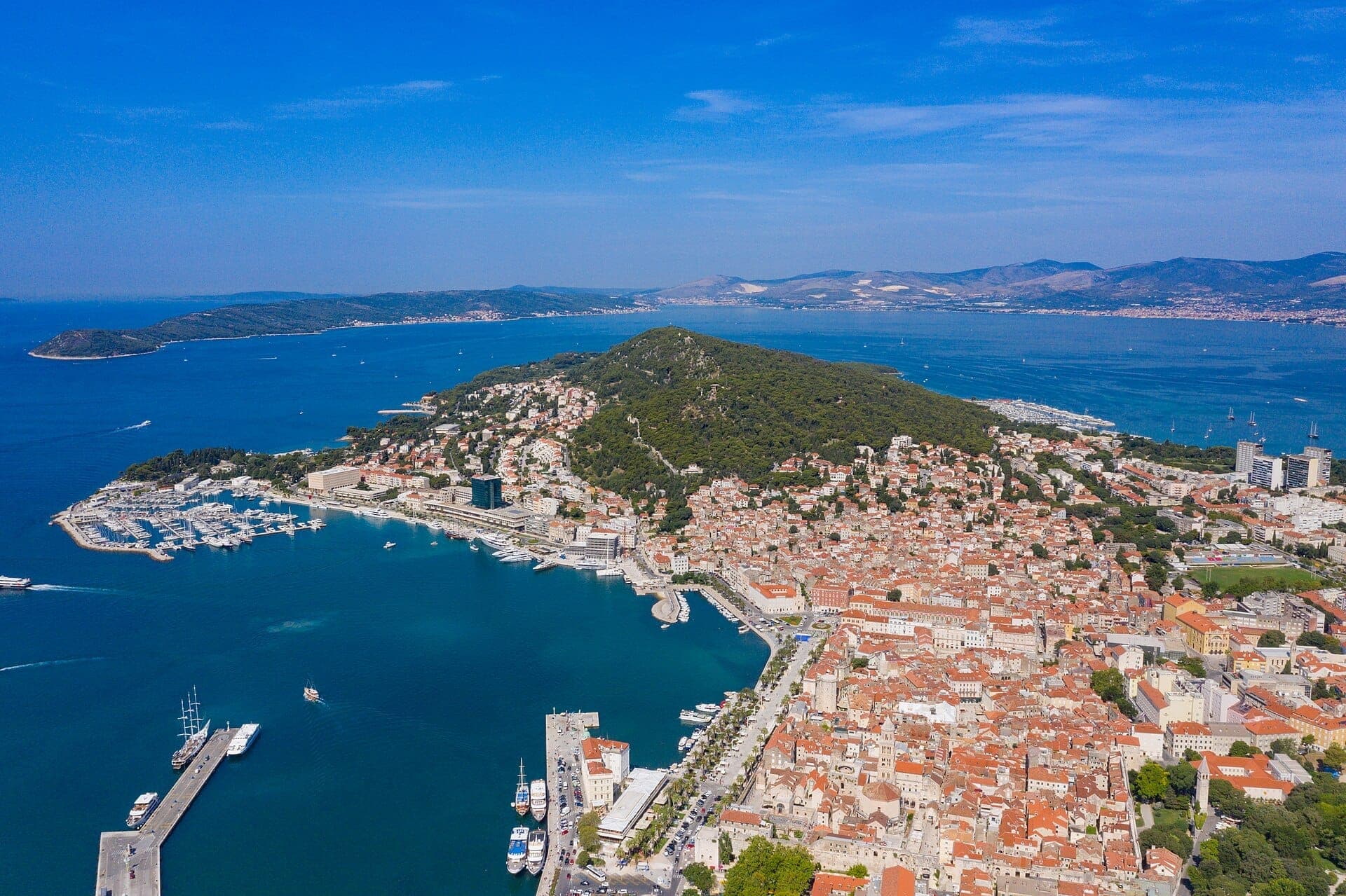 Vista aerea di Marjan Hill e della Penisola di Marjan, con folta foresta verde, il porto turistico di Split, edifici storici e le brillanti acque blu dell'Adriatico sotto un cielo sereno.