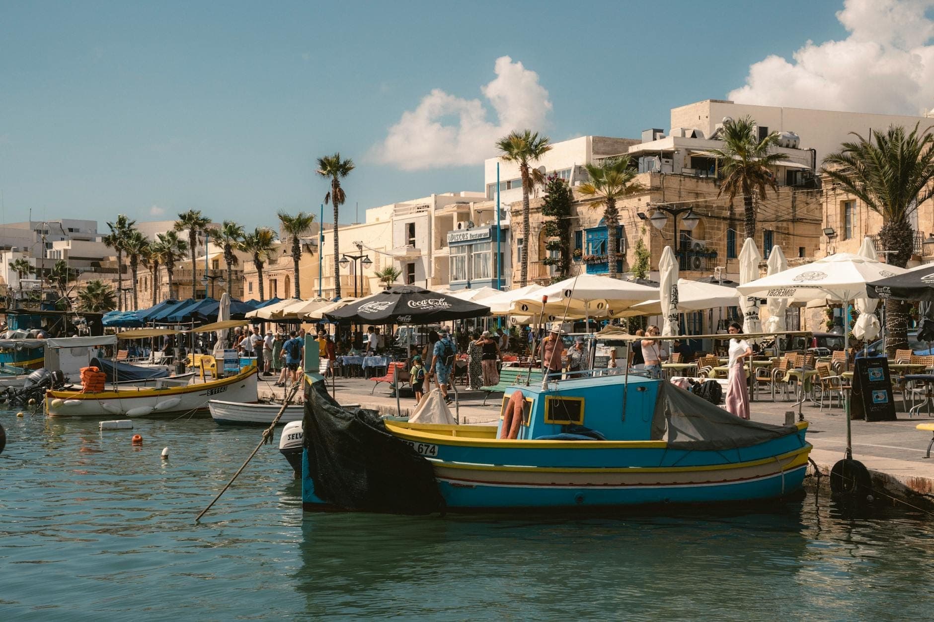 Traditionelle, bunt bemalte Fischerboote liegen an der Uferpromenade von Marsaxlokk, im Hintergrund geschäftige Marktstände, Cafés und Palmen.