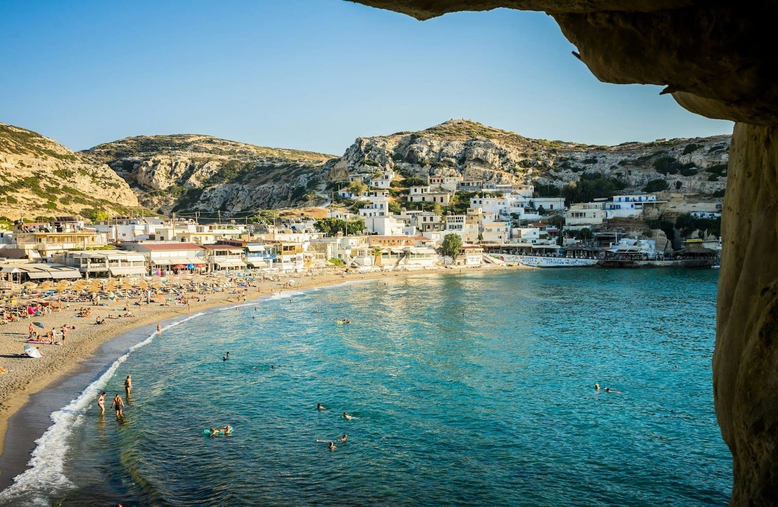Matala Beach mit türkisfarbenem Wasser, Schwimmern, Sandstrand, Sonnenschirmen und den berühmten Klippenhöhlen – an einem strahlend sonnigen Tag aus einer der Höhlen heraus fotografiert.