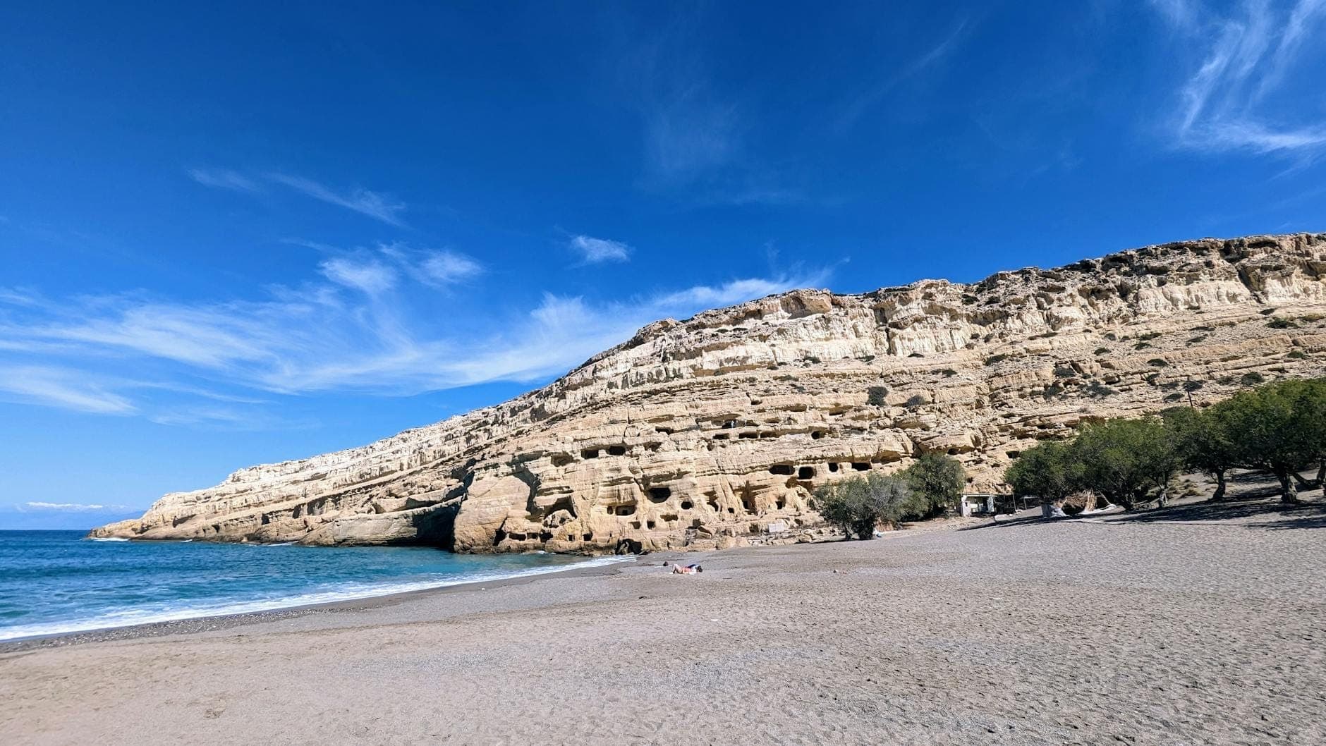 Breiter Sandstrand unterhalb der ikonischen Höhlen von Matala, in gelben Sandsteinfelsen gehauen, mit klarem Himmel, türkisblauem Meer und einigen Strandbesuchern.