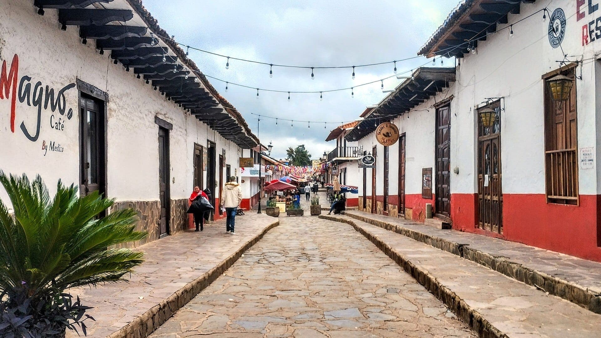 ถนนสายหลักในเมือง Mazamitla ที่ปูด้วยหินกรวด มีอาคารสไตล์โคโลเนียลสีขาวและสีแดง ไฟประดับห้อยอยู่เหนือศีรษะ และผู้คนเดินอยู่ไม่กี่คนในวันที่มีเมฆครึ้ม