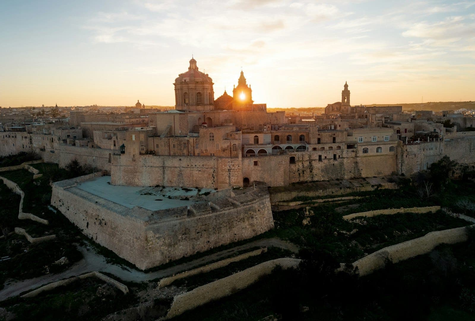 Luftaufnahme von Mdinas antiken Festungsmauern und der Kathedrale bei Sonnenuntergang – goldenes Licht fällt auf die historischen Steingebäude auf dem Hügel.