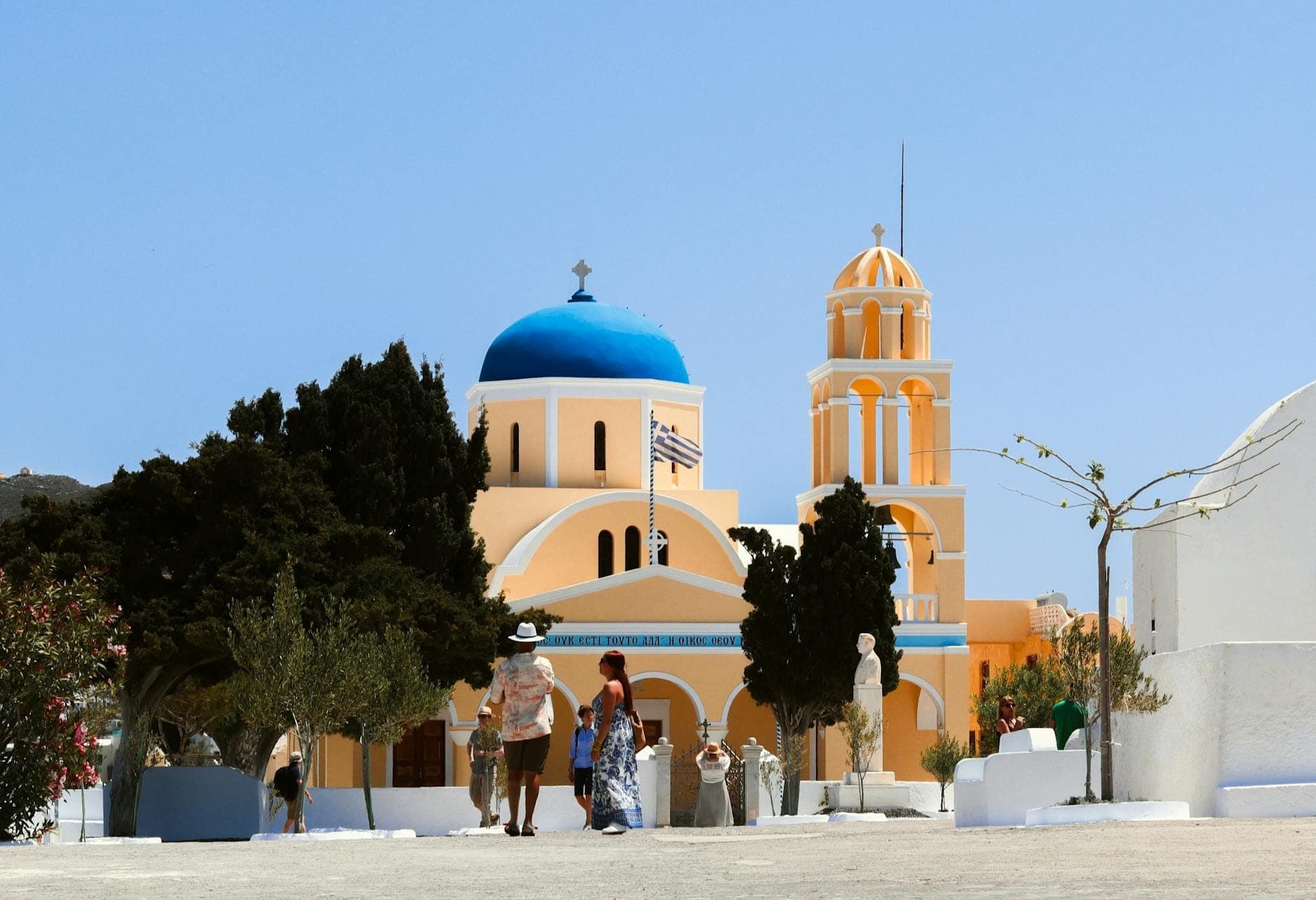 Traditionelle Kirche mit blauer Kuppel und Glockenturm im Dorf Megalochori auf Santorin, mit Spaziergängern auf einem sonnenbeschienenen, ruhigen Platz und weiß getünchten Häusern ringsum.