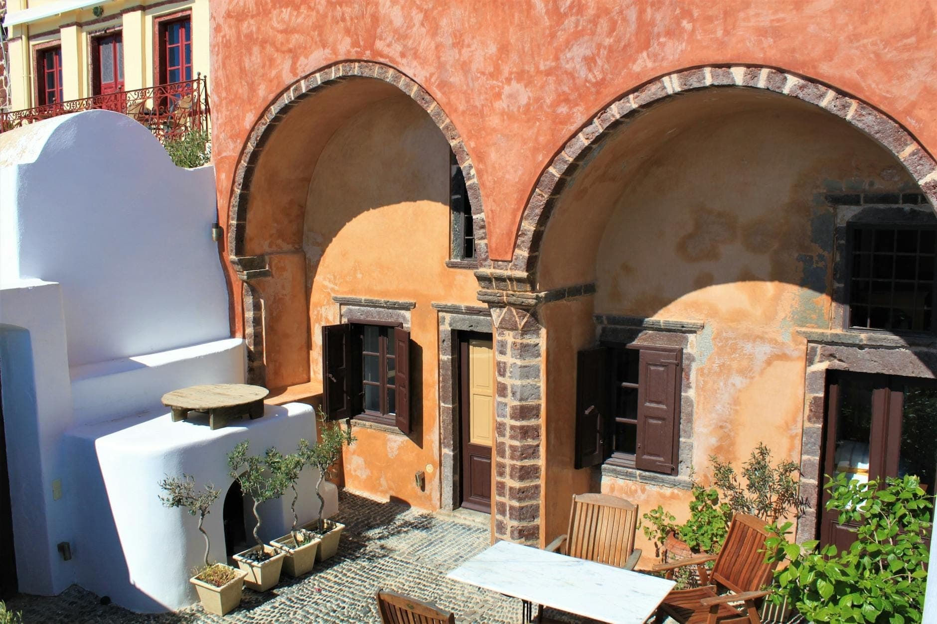 Sonnenbeschienene Fassade eines historischen ockerfarbenen Herrenhauses auf Santorin mit Rundbogentüren, Steindetails und einem kleinen Innenhof mit Tischen und Topfpflanzen.