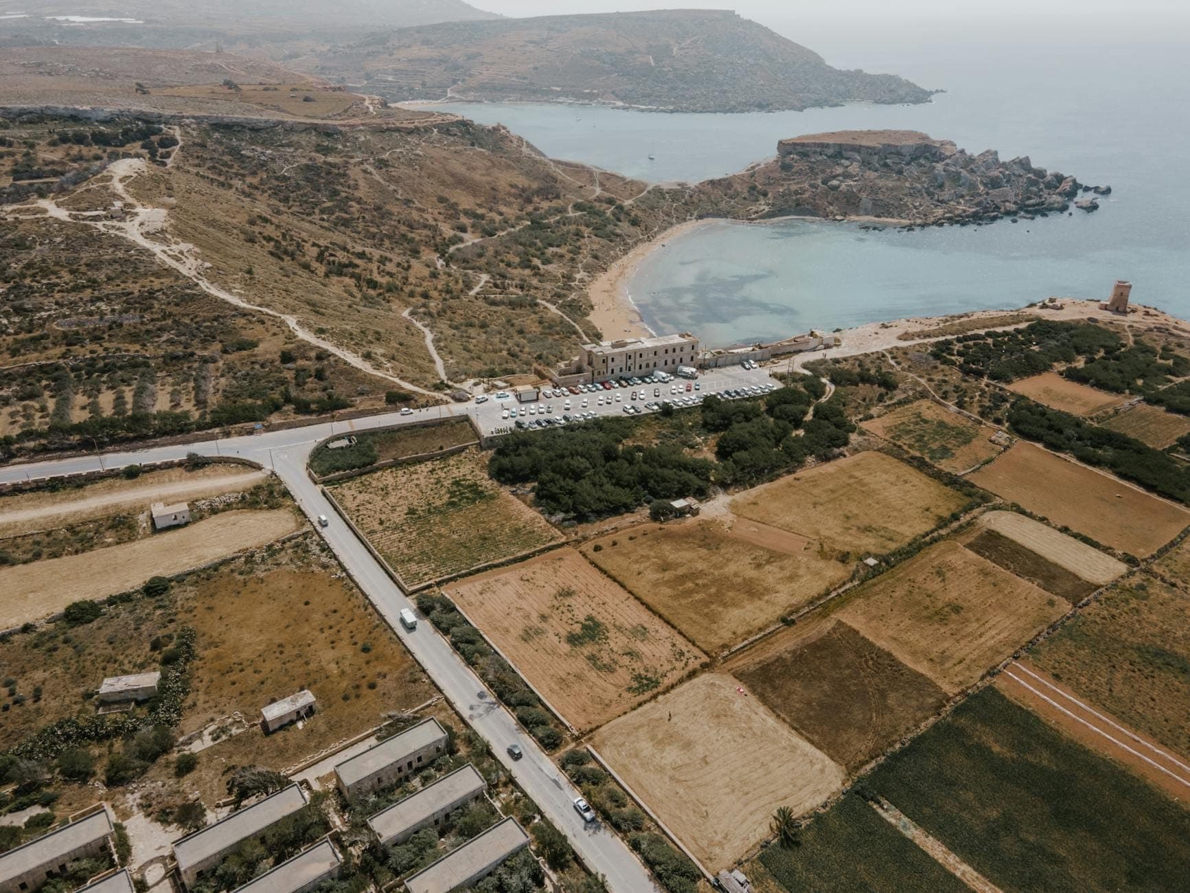 Luftaufnahme der Mellieħa Bay mit einem langen Sandstrand, ruhigem blauem Wasser, dem angrenzenden Naturschutzgebiet und landwirtschaftlichen Feldern entlang der maltesischen Küste.