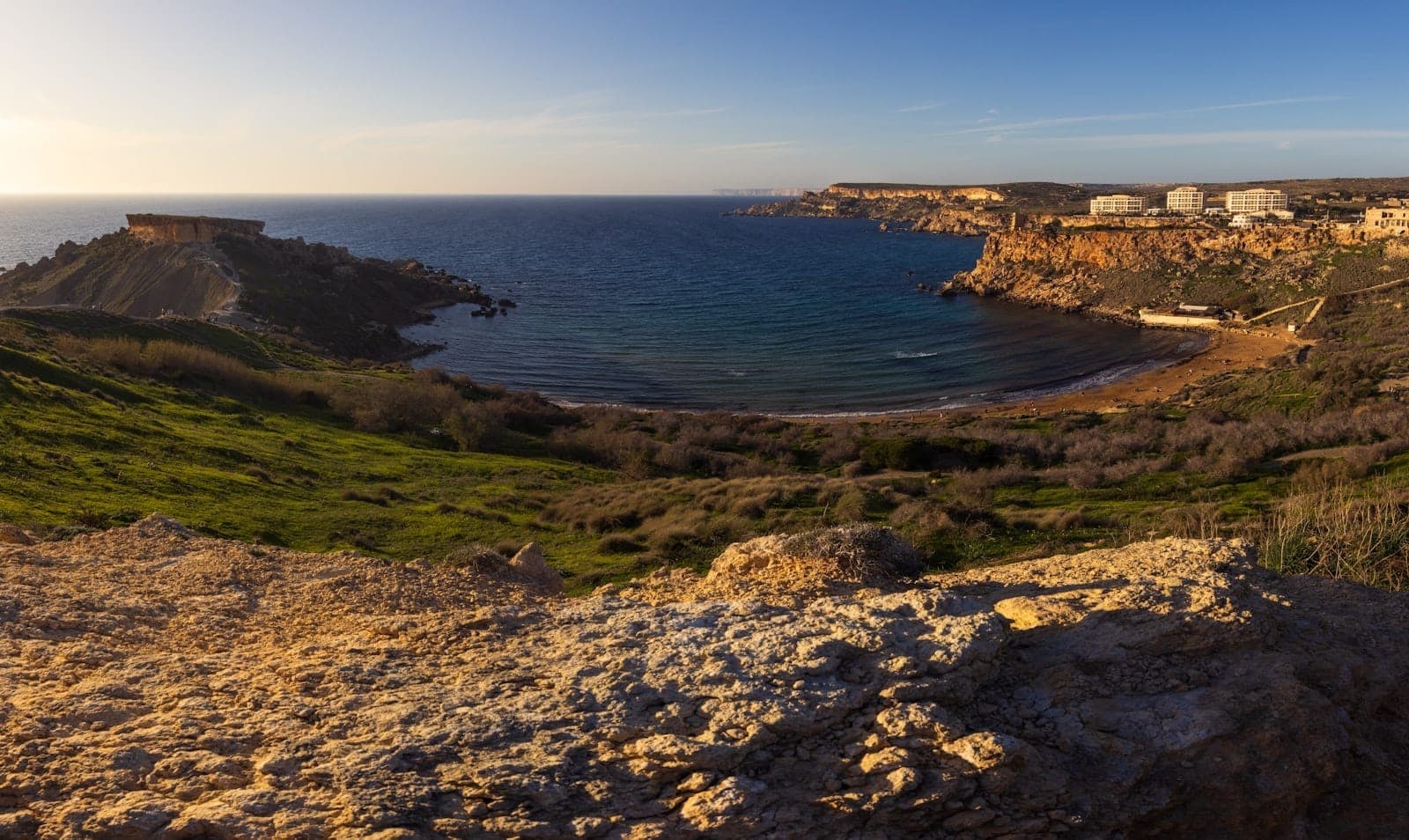 Weitläufige Landschaftsaufnahme der Mellieħa-Bucht bei Sonnenuntergang mit Klippen, Sandstrand, türkisfarbenem Meer und grünen Hügeln unter klarem Himmel.
