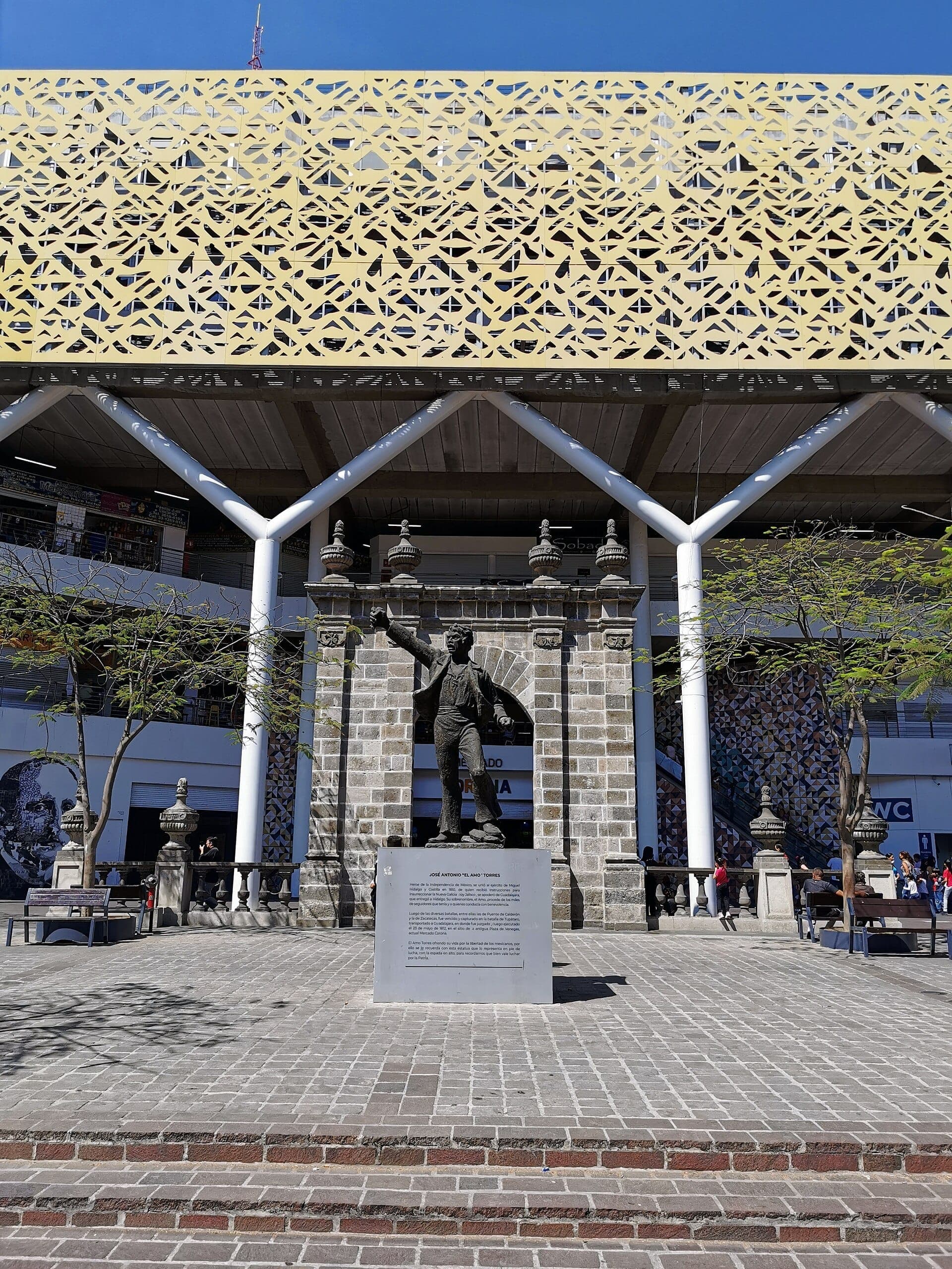 มุมมองด้านหน้าของ Mercado Corona ในเมือง Guadalajara โดดเด่นด้วยด้านหน้าอาคารสมัยใหม่ รูปปั้นกลางแจ้ง และลานกลางแจ้งในวันที่แดดจ้าและท้องฟ้าแจ่มใส