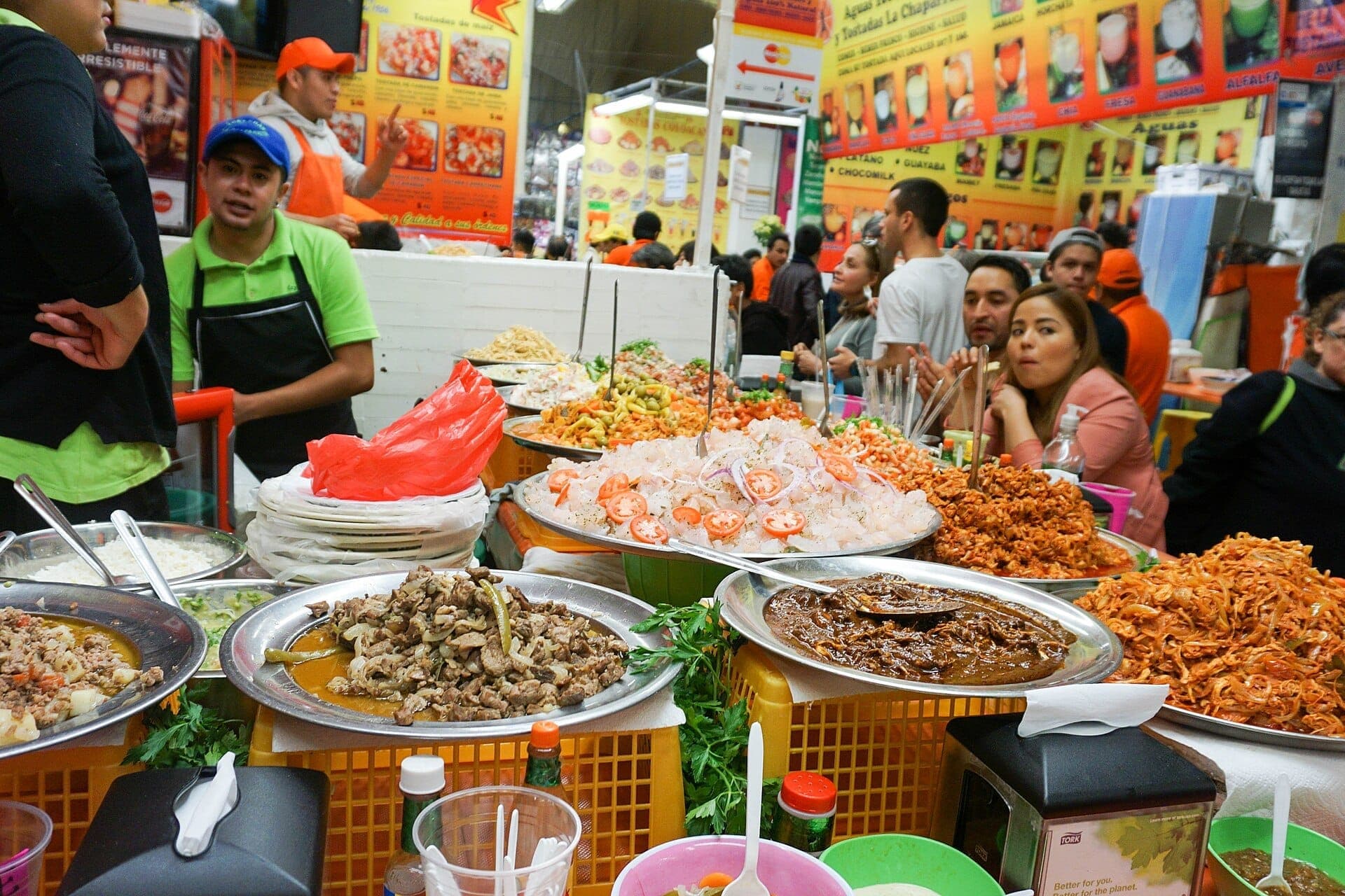 แผงอาหารที่คึกคักใน Mercado de Coyoacán พร้อมอาหารหลากสีสัน เจ้าหน้าที่สวมผ้ากันเปื้อน และฝูงชนที่สนุกสนานกับอาหารประเพณีเม็กซิกัน