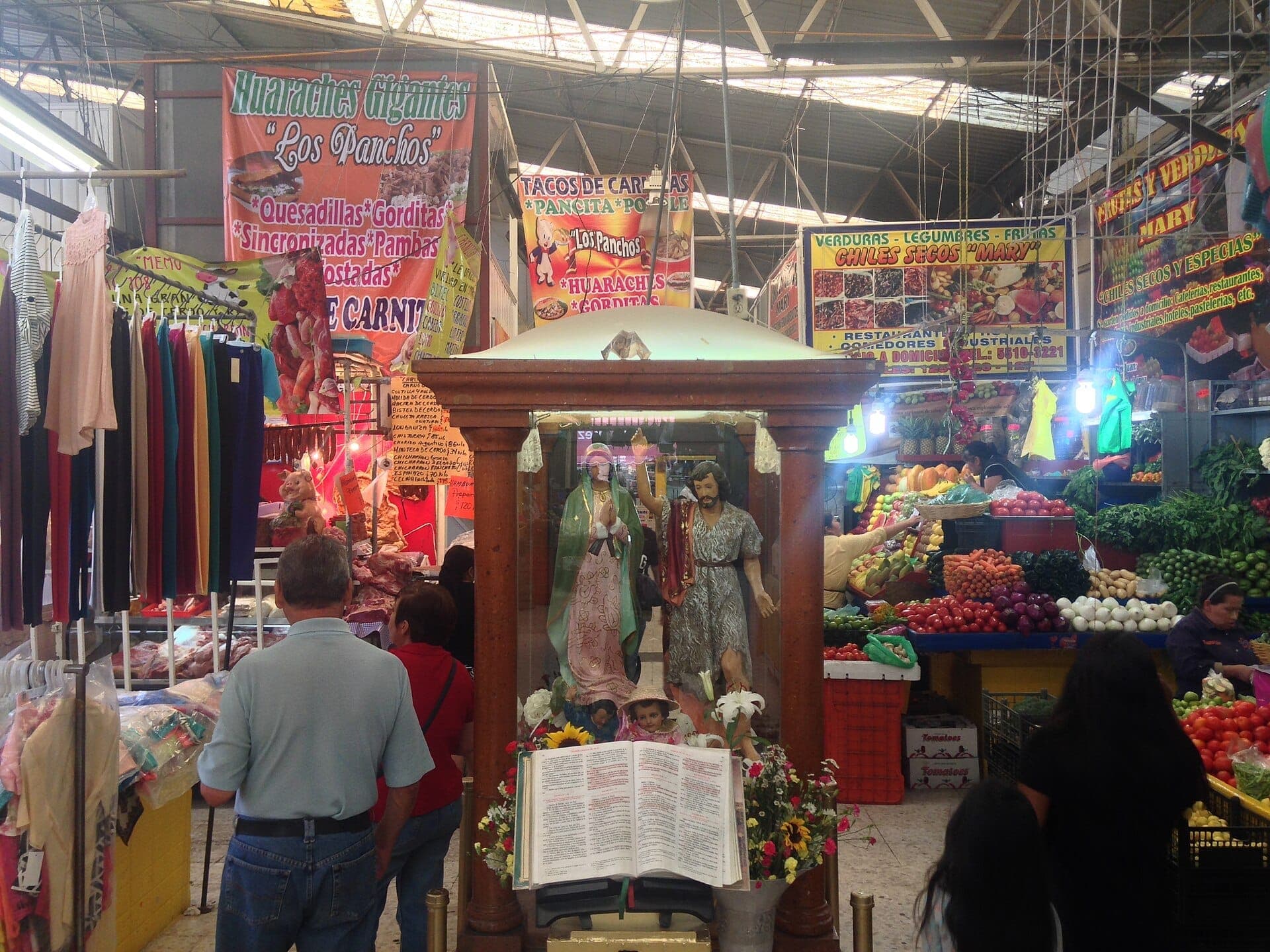 ภายในตลาด Mercado de San Juan ที่คึกคักพร้อมแผงขายสินค้าสีสันสดใส รูปปั้นทางศาสนา เสื้อผ้าแขวน และนักช้อปใต้แสงไฟฟลูออเรสเซนต์