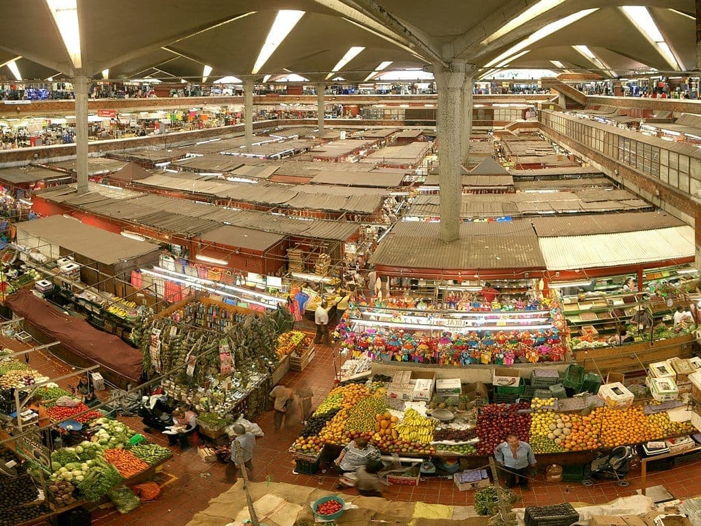 วิวภายใน Mercado San Juan de Dios แสดงให้เห็นแผงขายสินค้าเรียงเป็นแถว สินค้าหลากสีสัน และผู้คนที่กำลังช้อปปิ้งใต้หลังคาสมัยใหม่ขนาดใหญ่