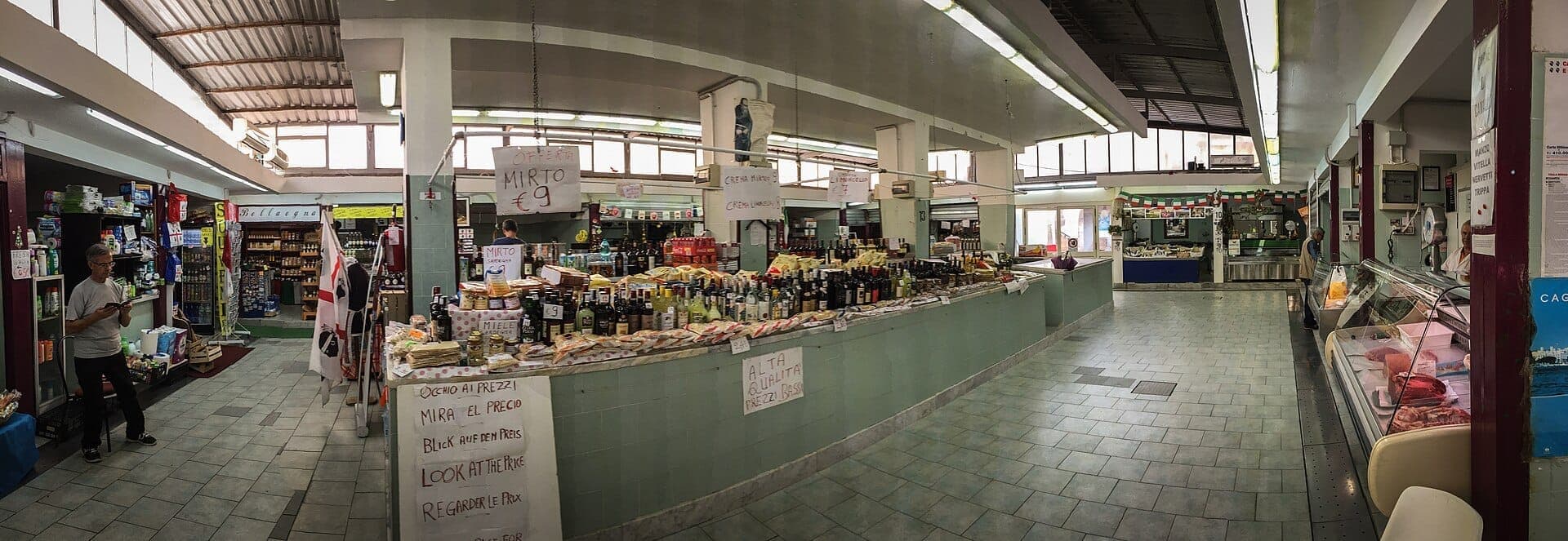 Vista amplia del interior del Mercato di San Benedetto, con varios puestos de comida, vendedores del mercado, estantes de productos embotellados y luz natural de grandes ventanas.