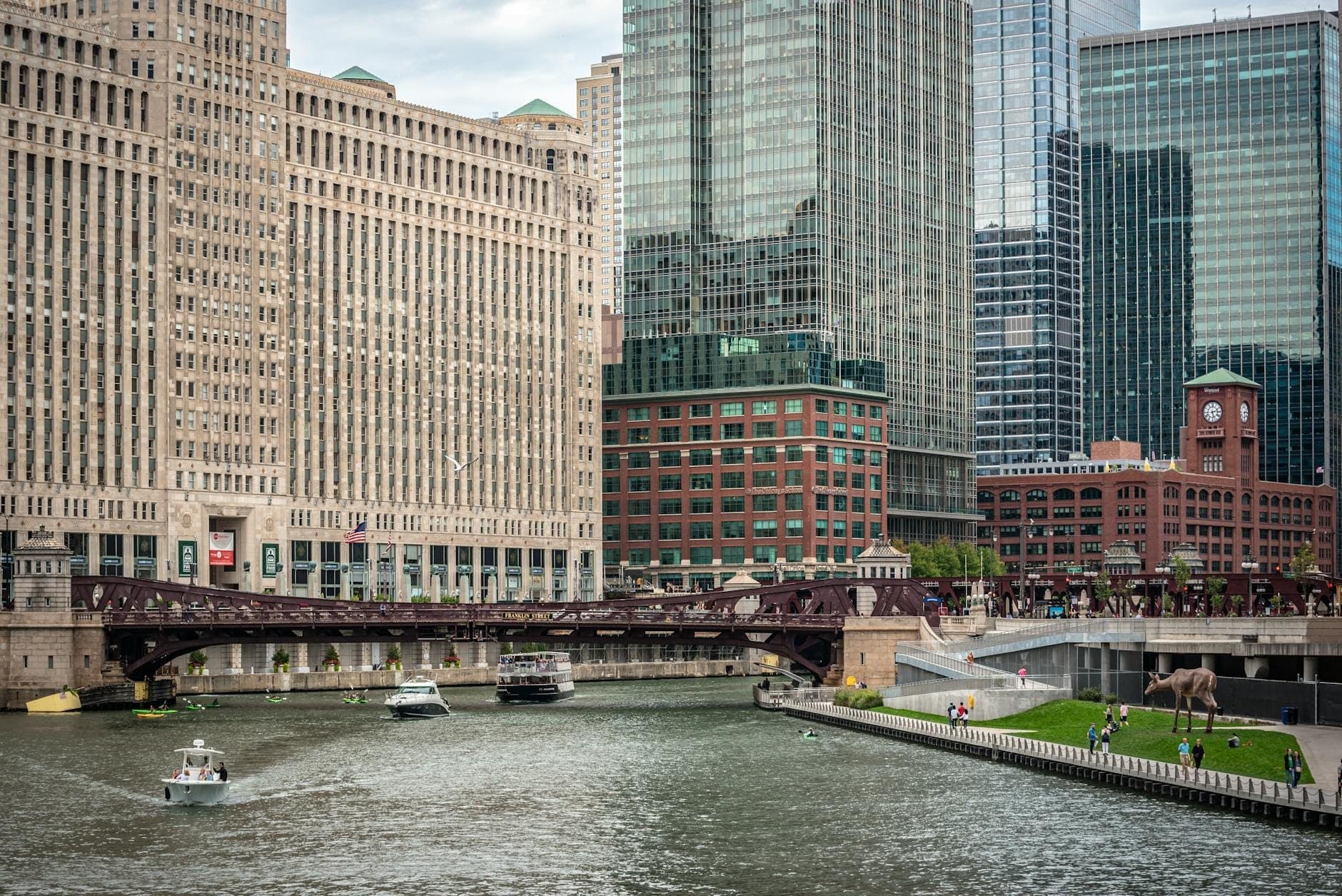 The Merchandise Mart building rises prominently along the Chicago River, with boats on the water and modern skyscrapers surrounding the landmark in downtown Chicago.