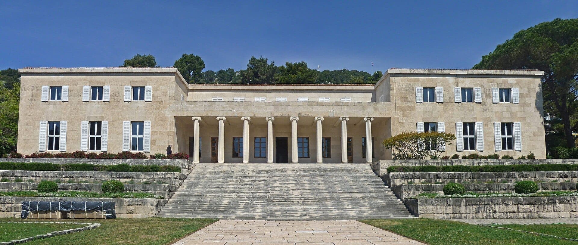 Vista frontale della Galleria Meštrović con la sua facciata neoclassica simmetrica, alte colonne, gradini in pietra e cespugli potati sotto un cielo azzurro limpido.