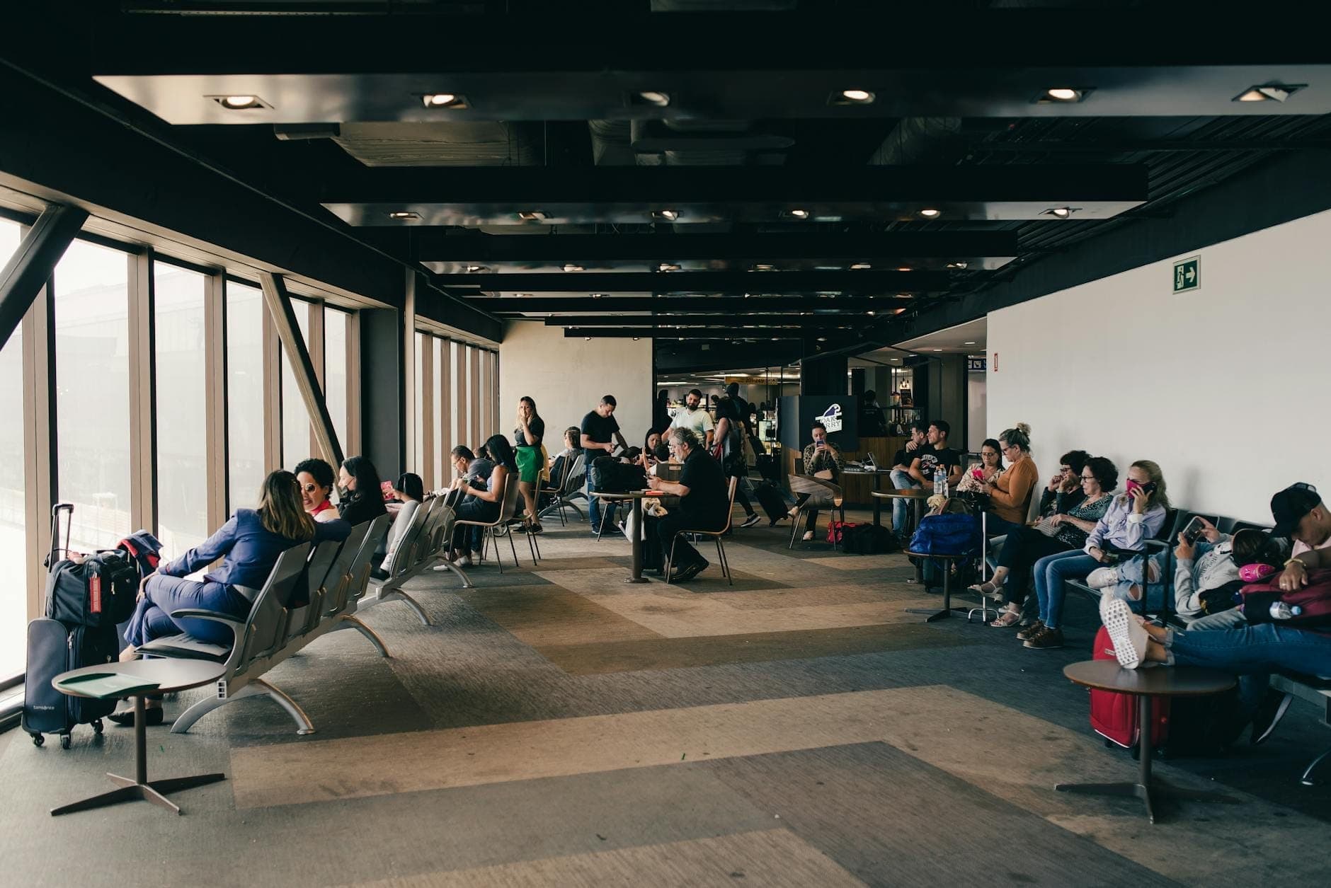 Travelers seated and waiting in a modern airport departure lounge area with large windows and visible airport seating.