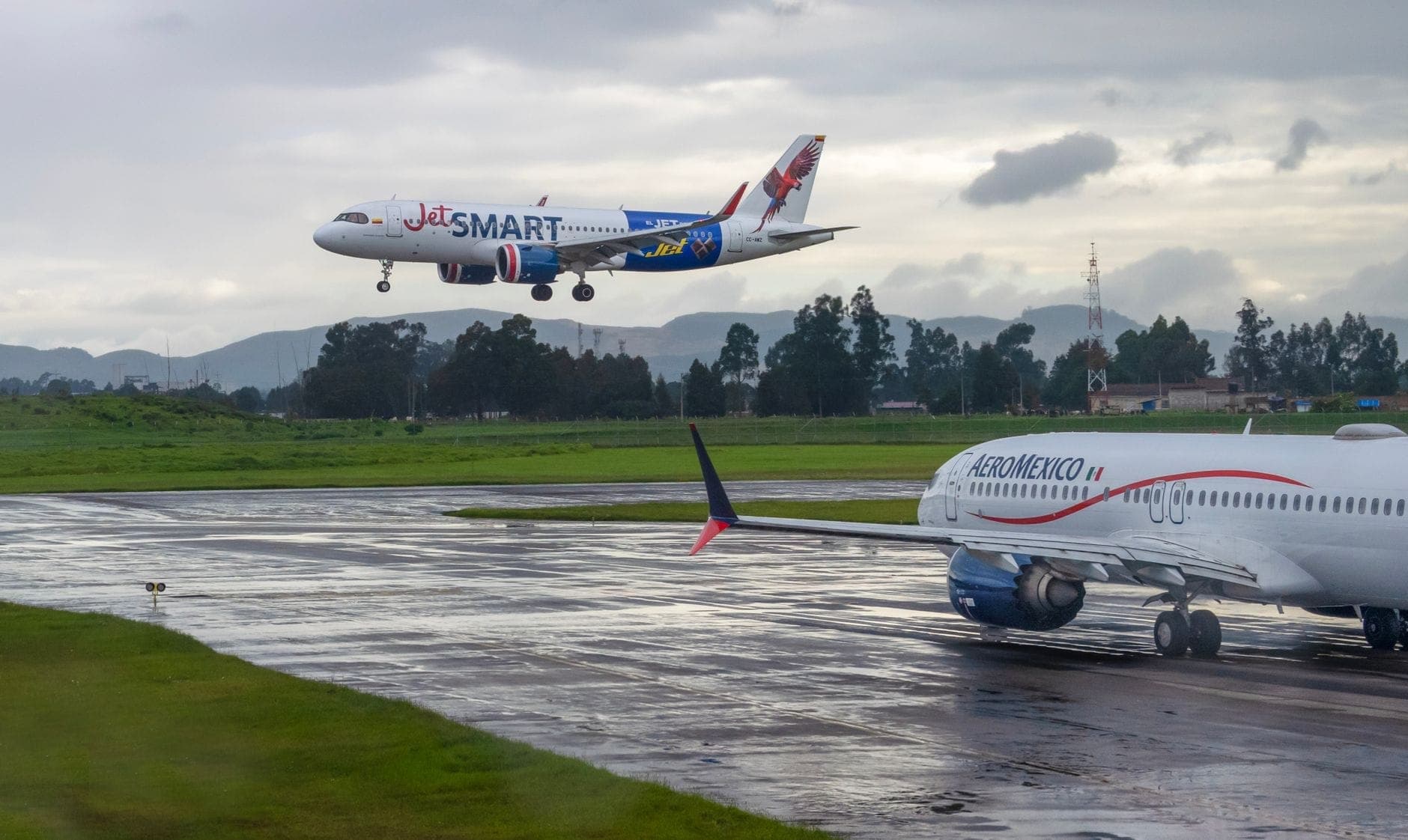Two commercial airplanes, one from JetSMART landing and one from Aeroméxico taxiing, on a wet runway with green surroundings and cloudy skies.
