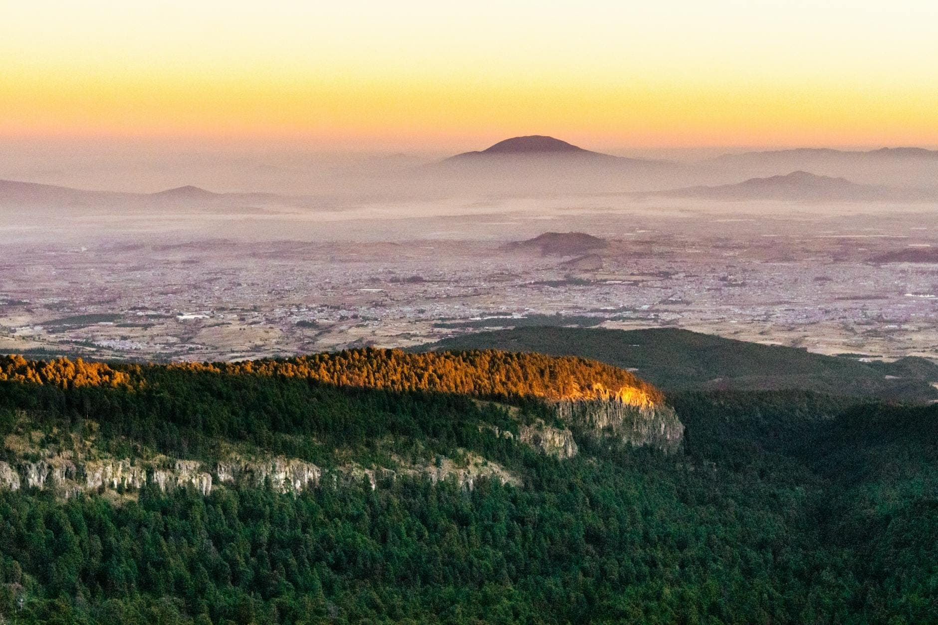 Vista panoramica dall'alto di Mexico City che si estende su un'ampia valle con montagne distanti e creste boscose all'alba.