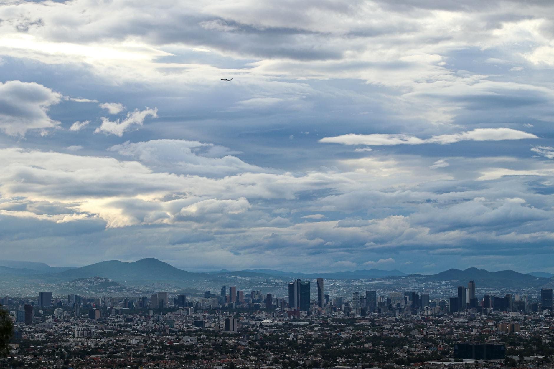 Ampia vista aerea di Città del Messico con edifici fitti, montagne in lontananza e un aereo in volo sotto un cielo spettacolare.