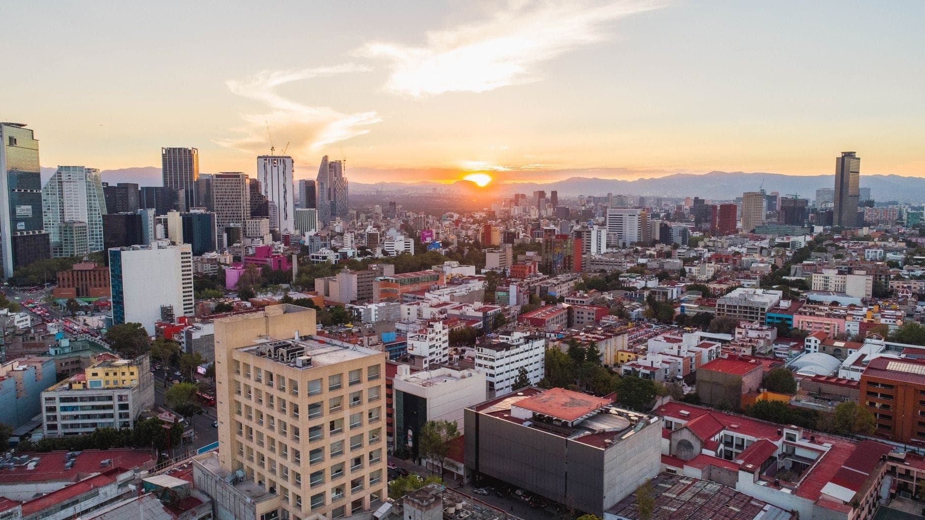 Vista aerea dello skyline di Città del Messico al tramonto con edifici moderni, aree residenziali e montagne sullo sfondo.