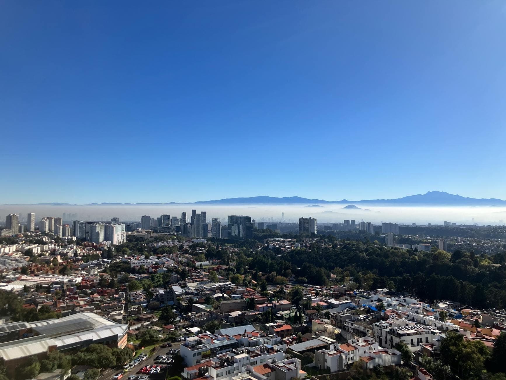 Vista nitida dello skyline di Città del Messico con edifici moderni, quartieri e montagne lontane sotto un cielo azzurro brillante e una leggera foschia.