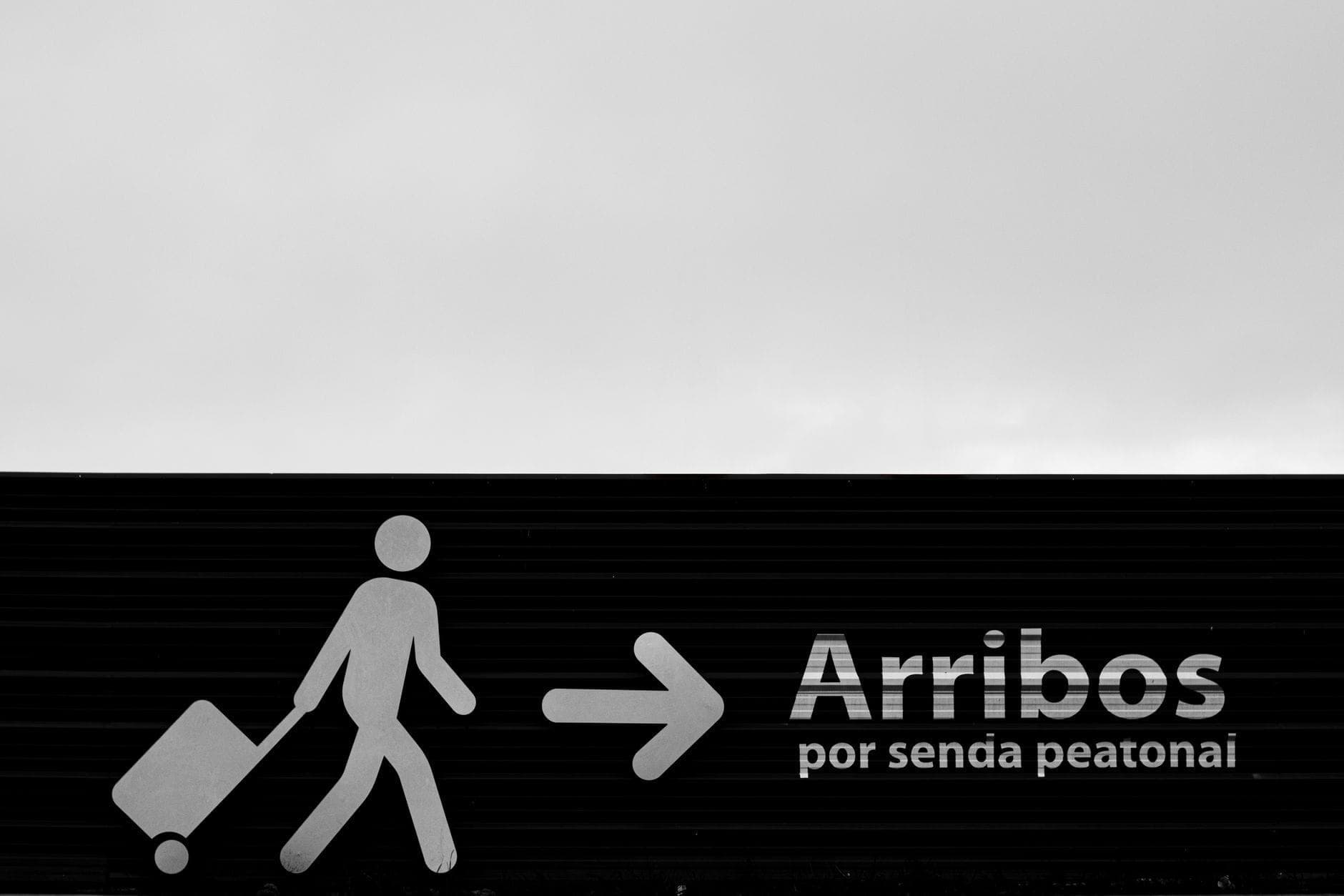 Letrero en blanco y negro en un aeropuerto que muestra a un viajero con maleta y la palabra 'Arribos' para llegadas.