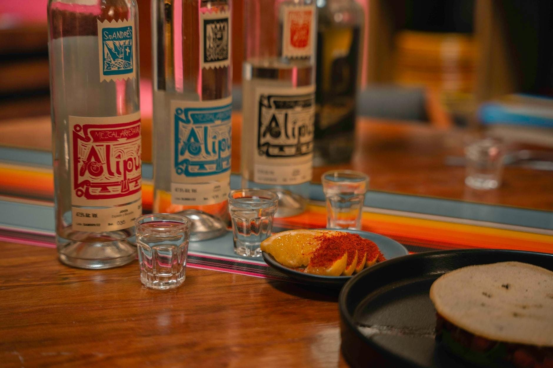 โต๊ะที่จัดวางด้วยขวด mezcal แก้วเล็ก ส้มหั่นแว่นโรยพริก และ taco แบบดั้งเดิมในร้านอาหารบรรยากาศอบอุ่น