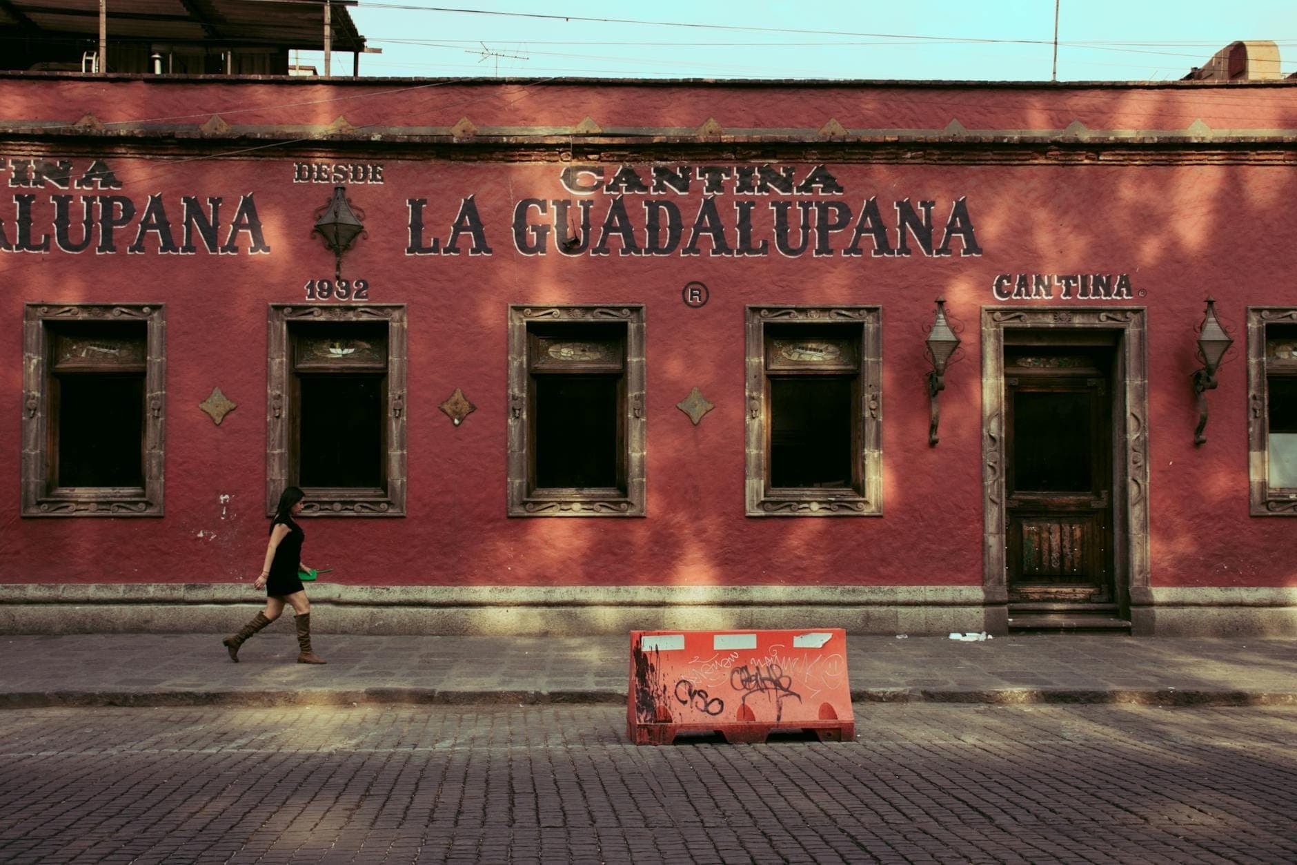 บาร์คันตินาแบบดั้งเดิม La Guadalupana ที่มีหน้าอาคารสีแดงใน Mexico City พร้อมผู้หญิงคนหนึ่งกำลังเดินผ่านถนนที่ปูด้วยหินกรวด