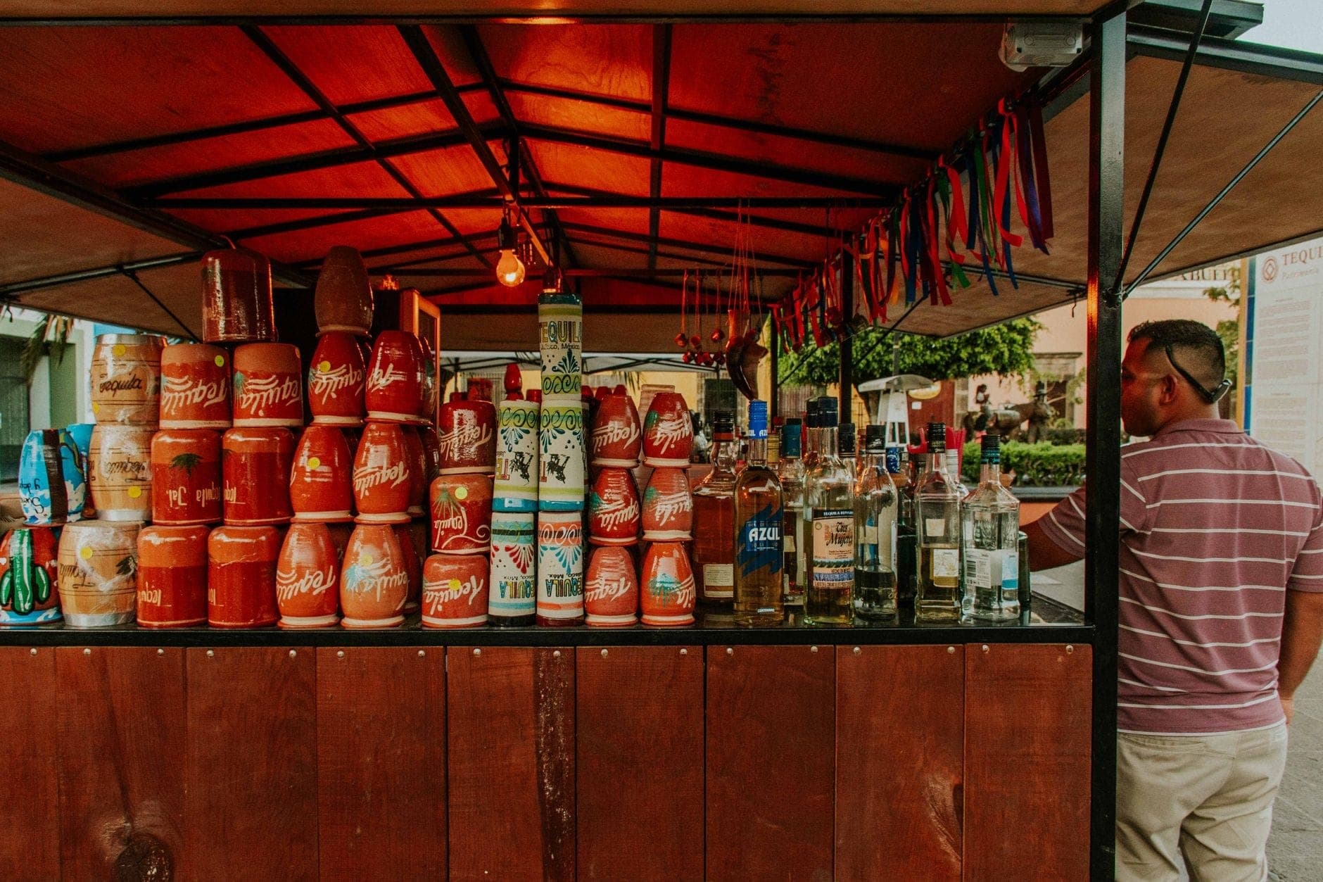 แผงตลาดไม้ใน Mexico City ที่มีขวดเมซคาลหลายขวดและถ้วยดินเหนียวแบบดั้งเดิมวางจัดแสดง ดูแลโดยชายสวมเสื้อลายทาง