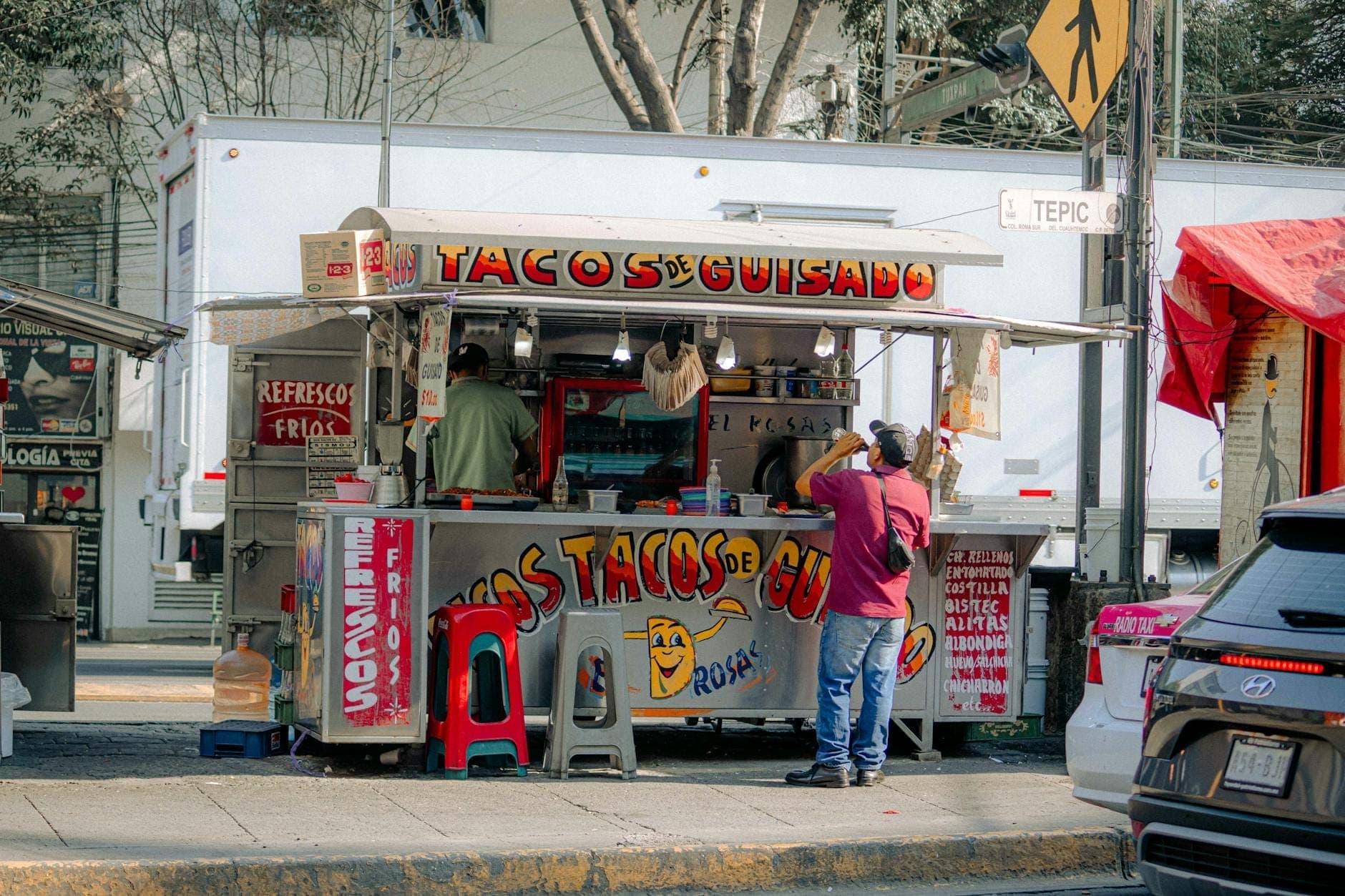 Street taco stand labeled 'Tacos de Guisado' with two people preparing food and colorful stools on a Mexico City sidewalk.